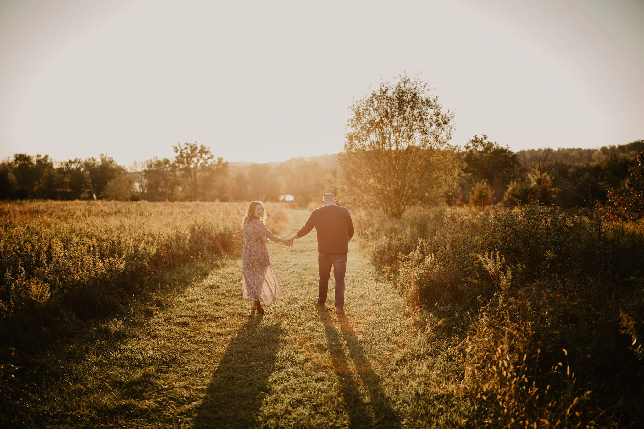 a couple (man and woman) are walking away from the camera into the sunset in a large golden field