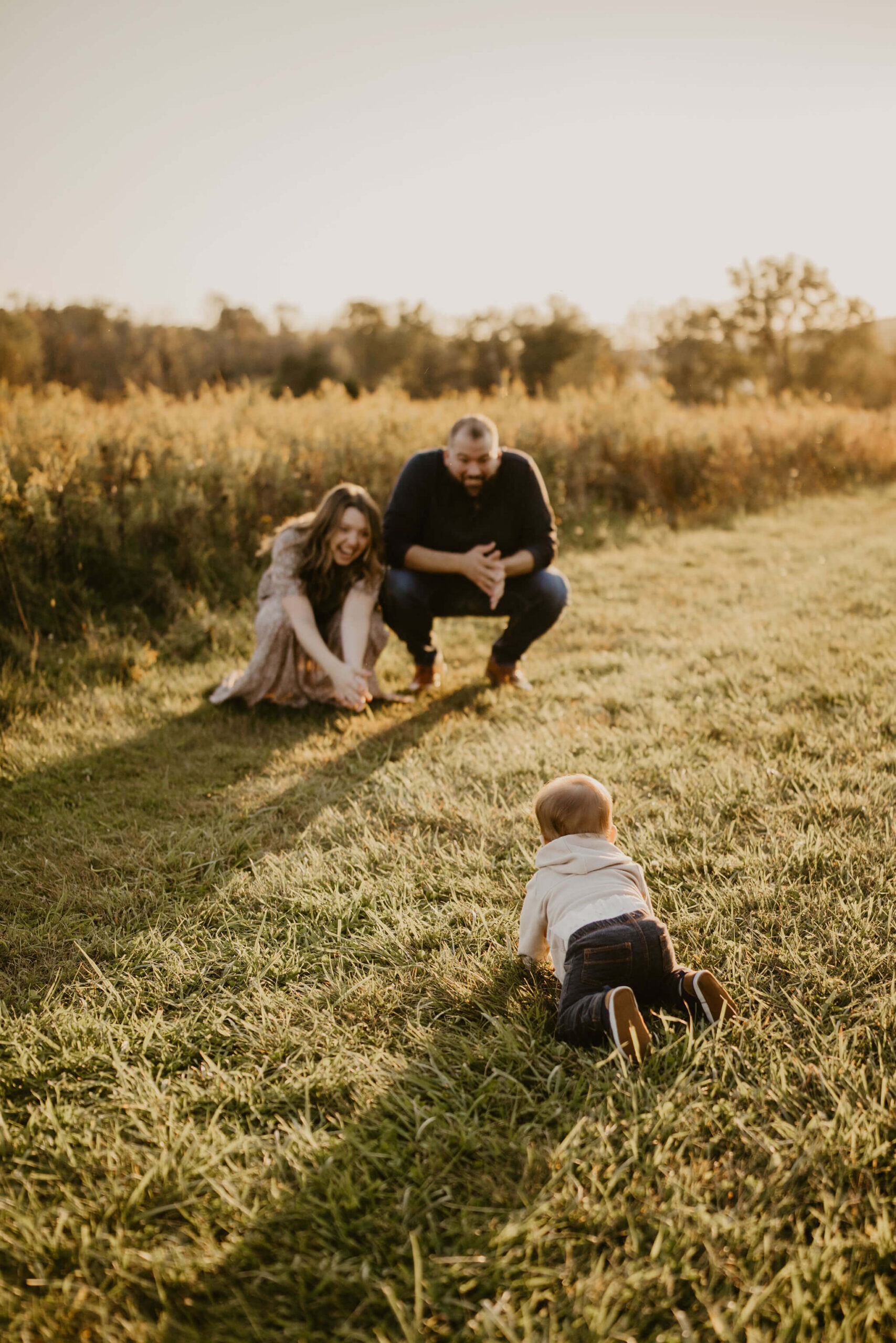 a baby in a field crawls towards his parents, who are crouched down to catch him