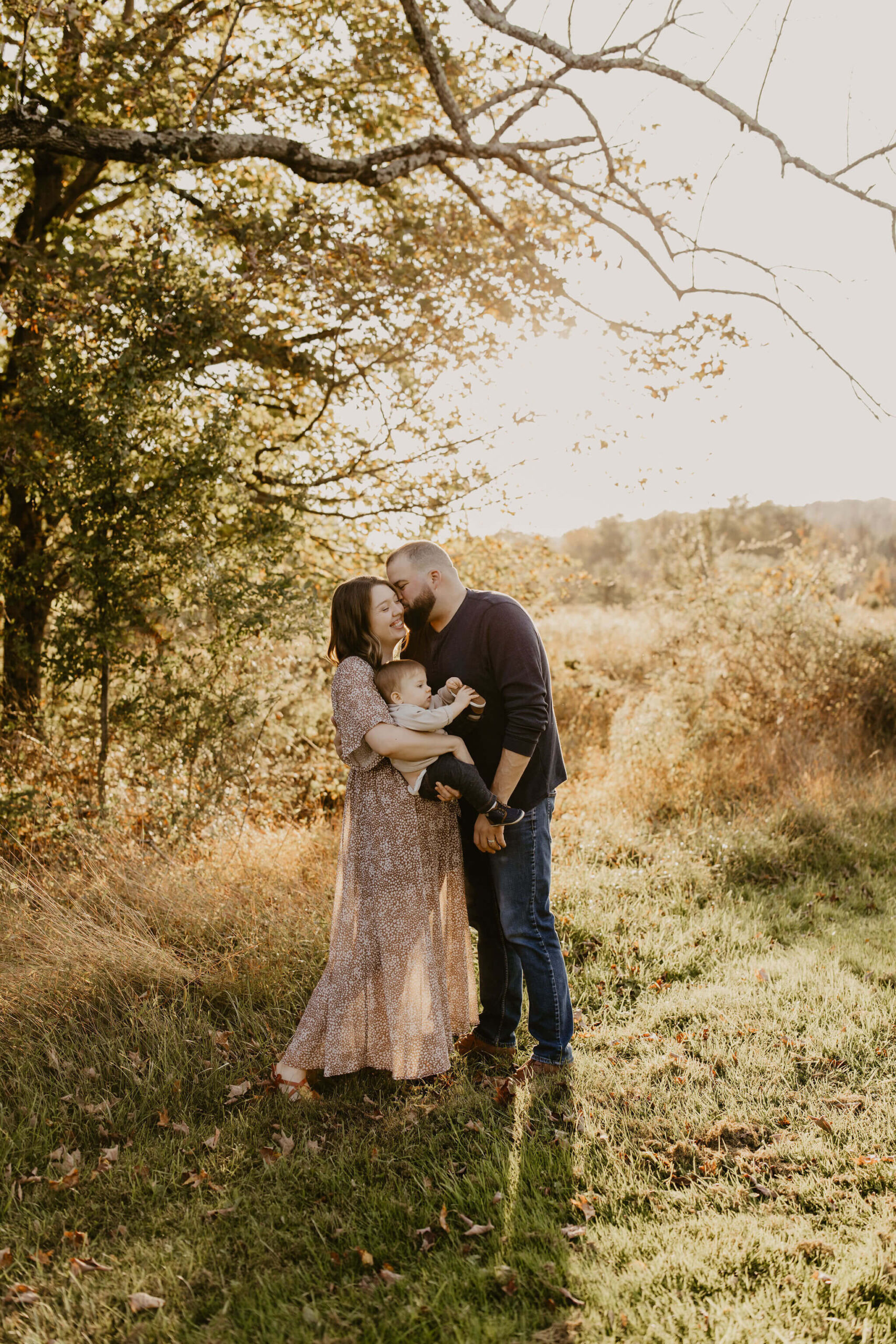 a father kissing his wife while she holds their young son, in a field with tall grass