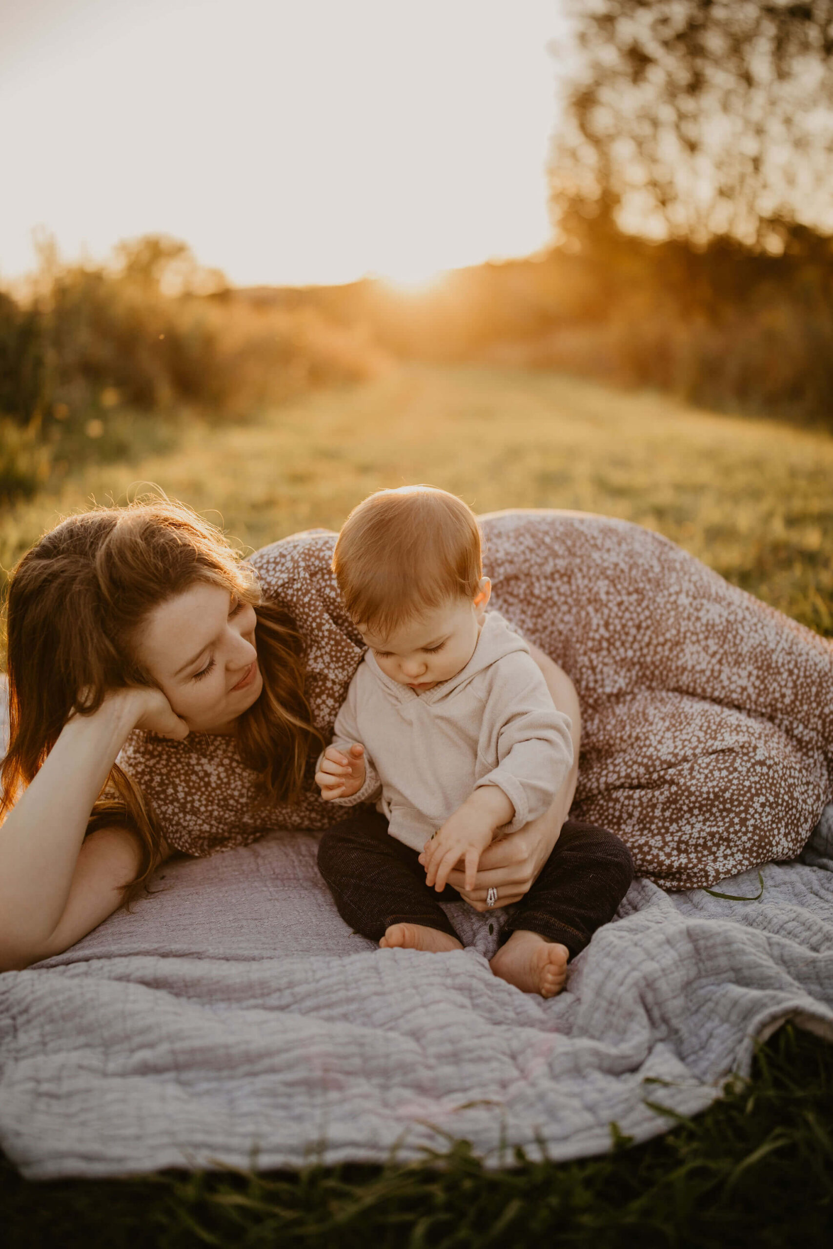 a mother and her son laying on a picnic blanket in a field at sunset