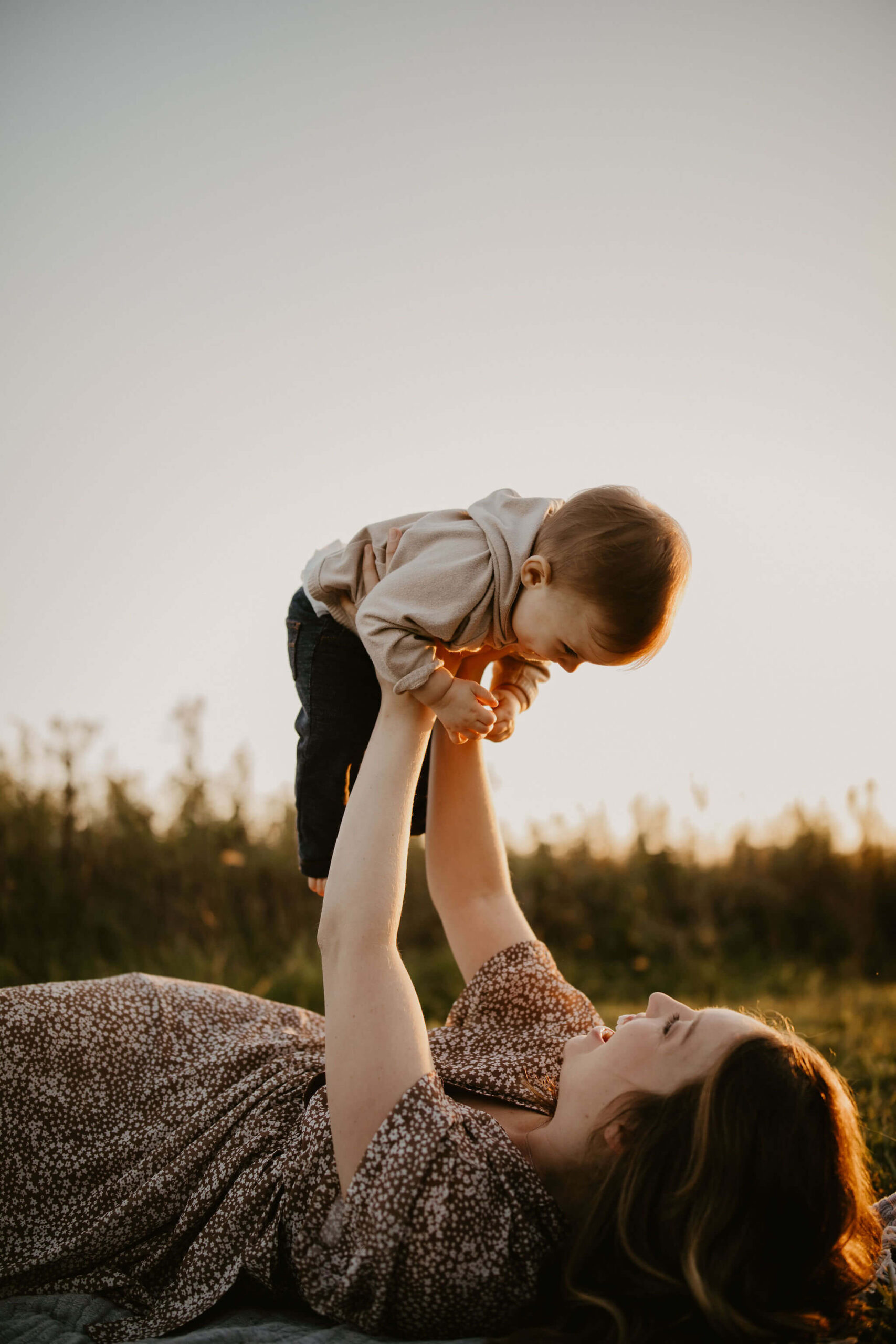 a mother and her son laying on a picnic blanket in a field at sunset. She is tossing him above her in the air and he is smiling down at her.