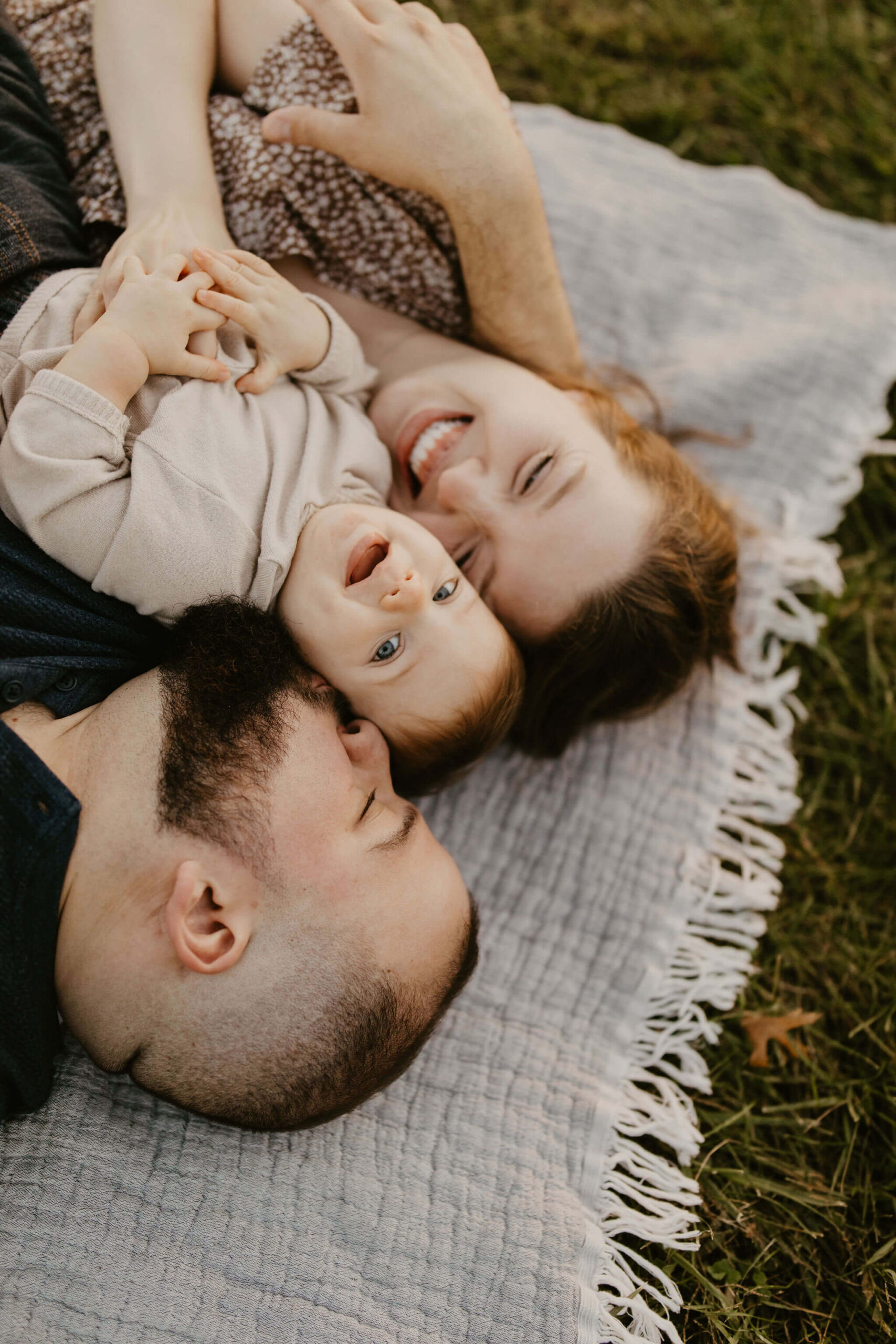 a family (father, mother, son) laying on a picnic blanket, cuddling together and laughing