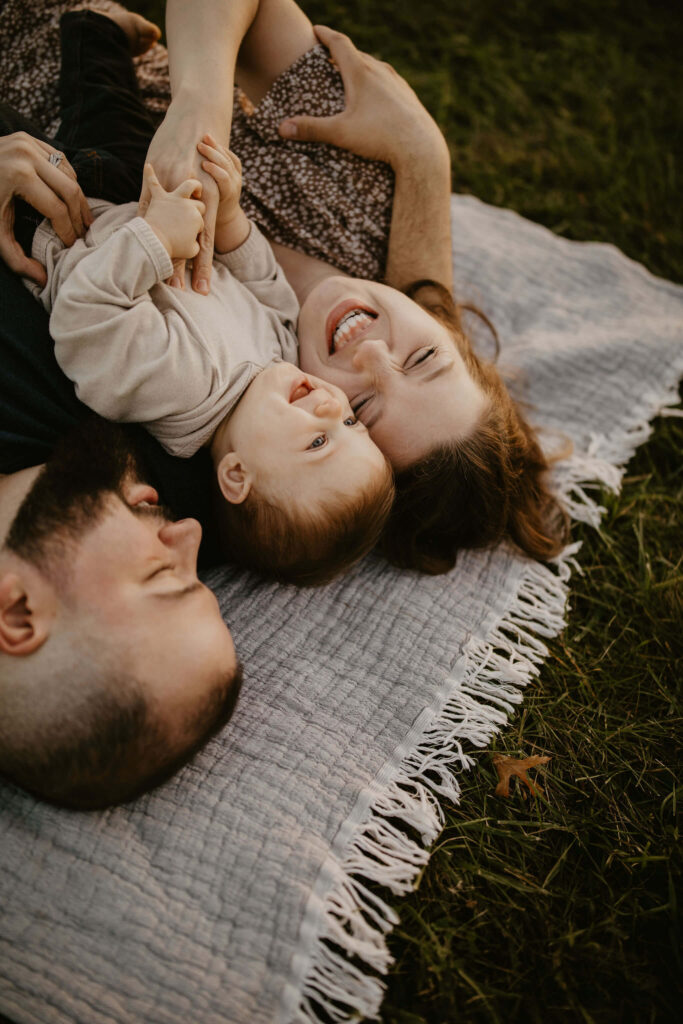 a family (father, mother, son) laying on a picnic blanket, cuddling together and laughing