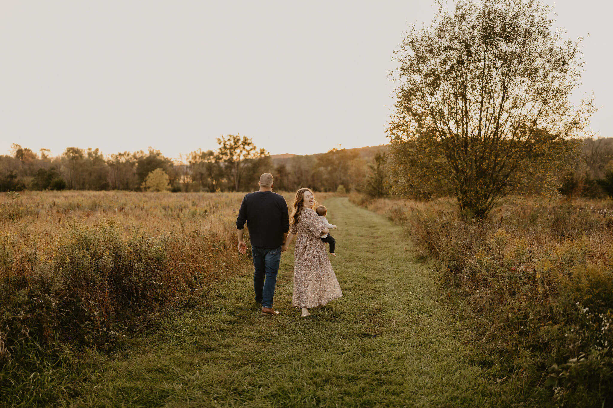 a family (father, mother, and son) walking away from the camera toward the sunset in a field during fall