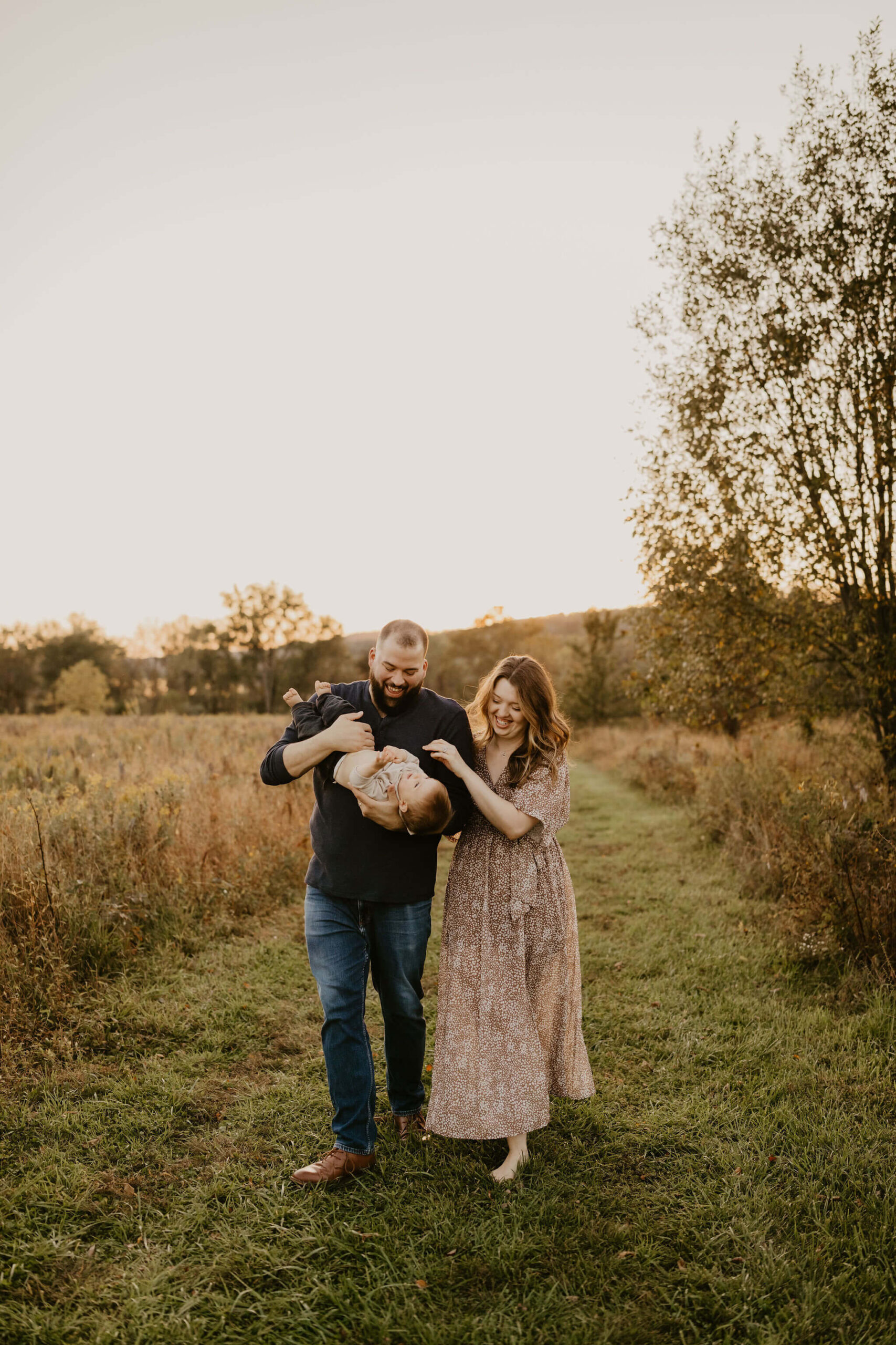 a family (mother, father, and son) walking toward the camera through a field at sunset. The son is being held upside down and giggling.