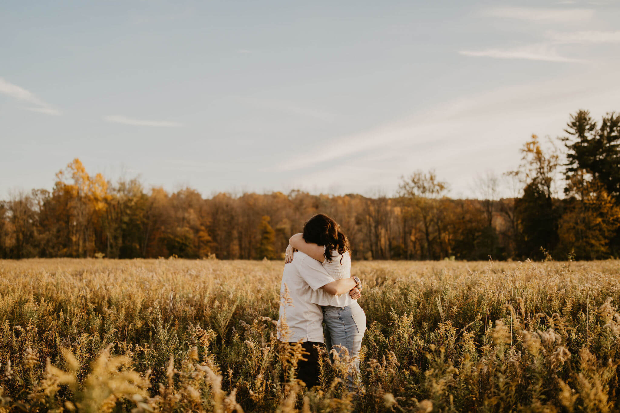 a boy and a girl, both with long brown wavy hair and white shirts, embracing in a golden field at sunset