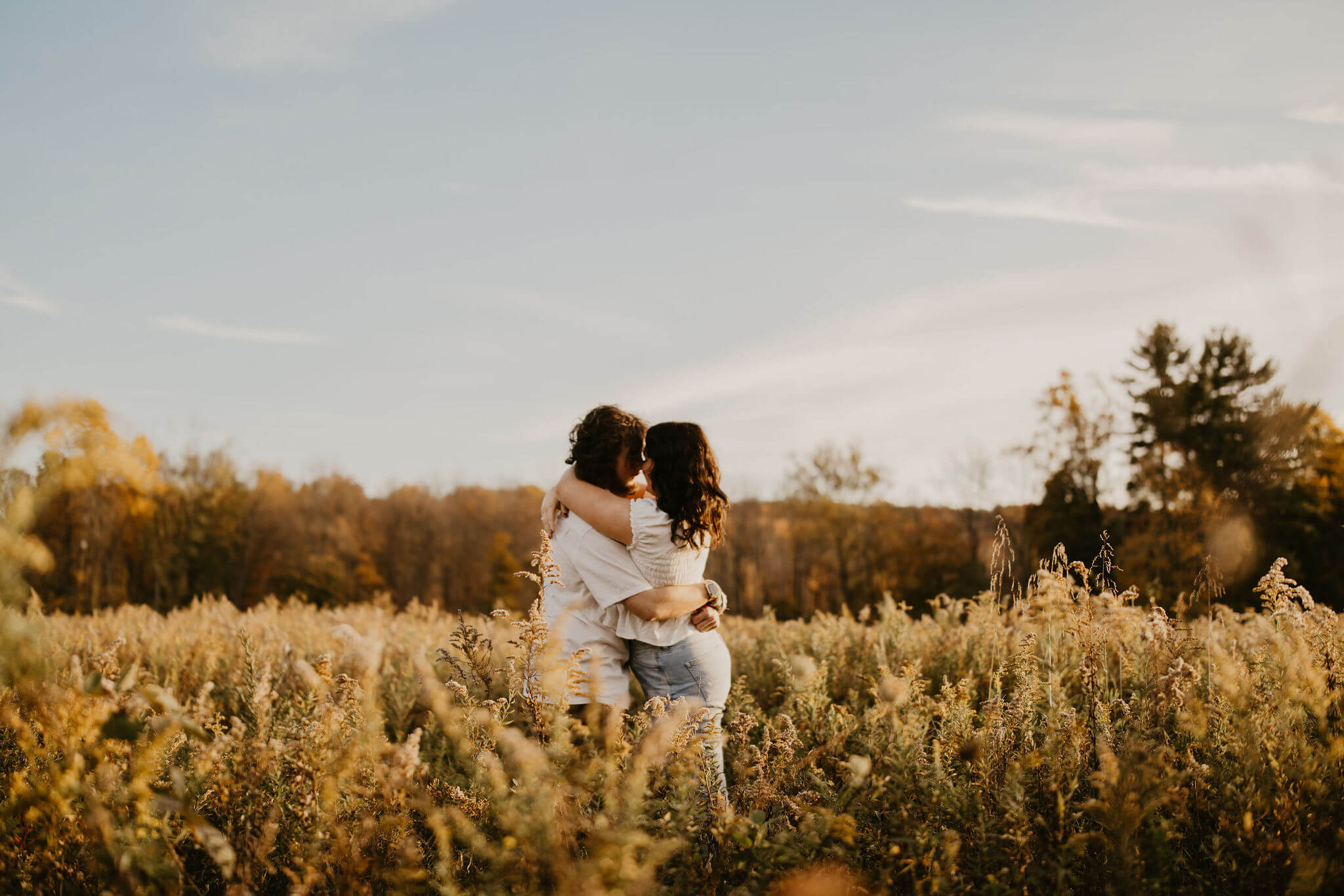 a couple (both brown hair with white shirts) embracing in a tall golden field at sunset