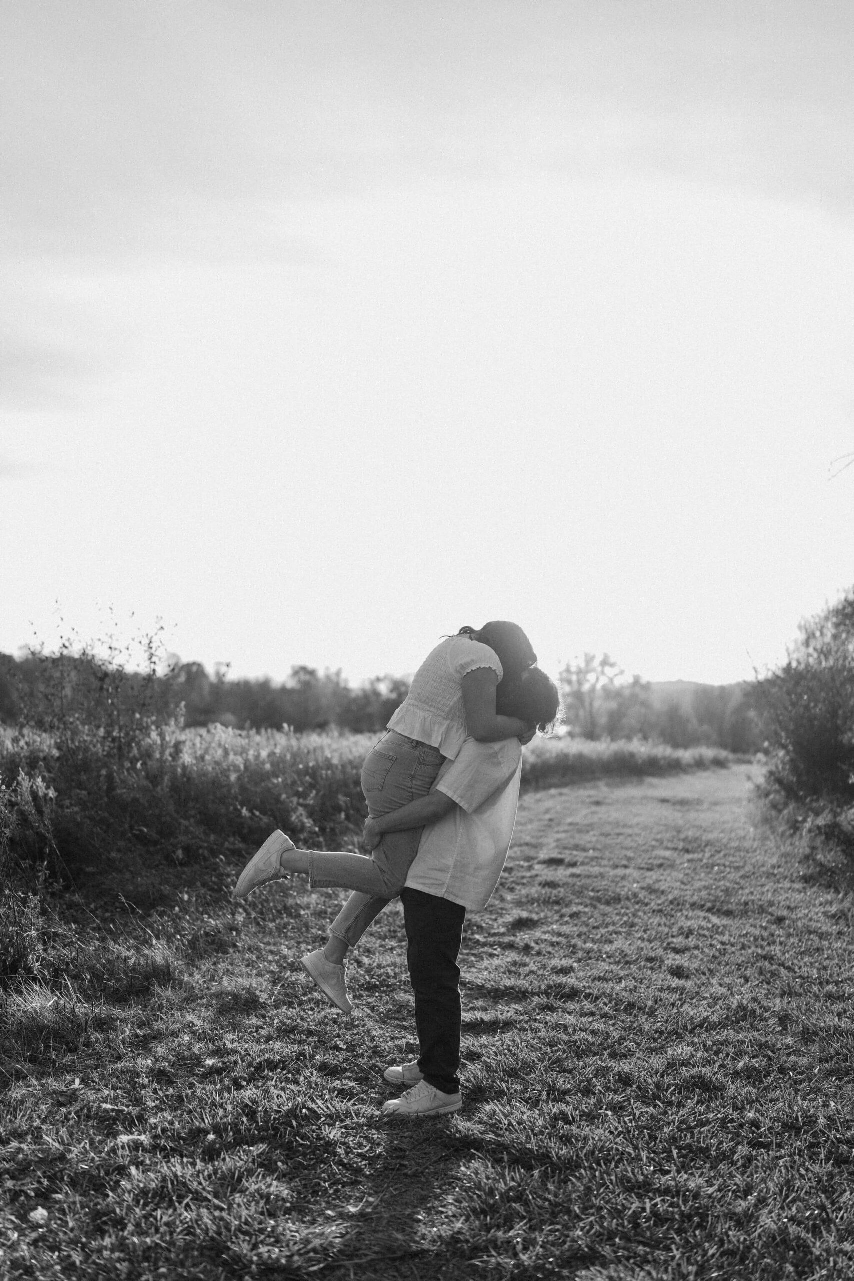 black and white image of a boy picking up his girlfriend and kissing her in a field