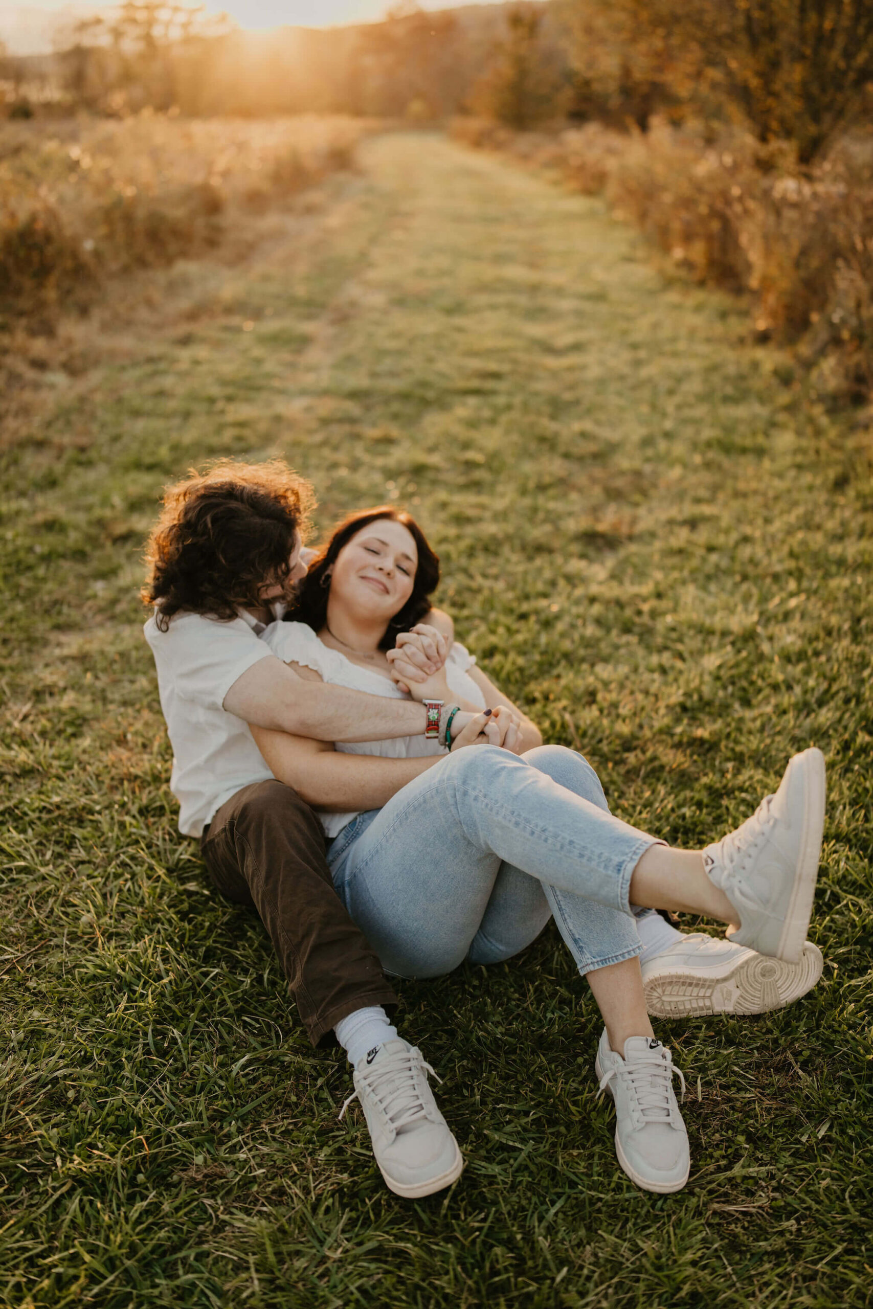 a couple cuddling in a field during golden hour. his arms are wrapped around her and she is smiling softly at the camera.