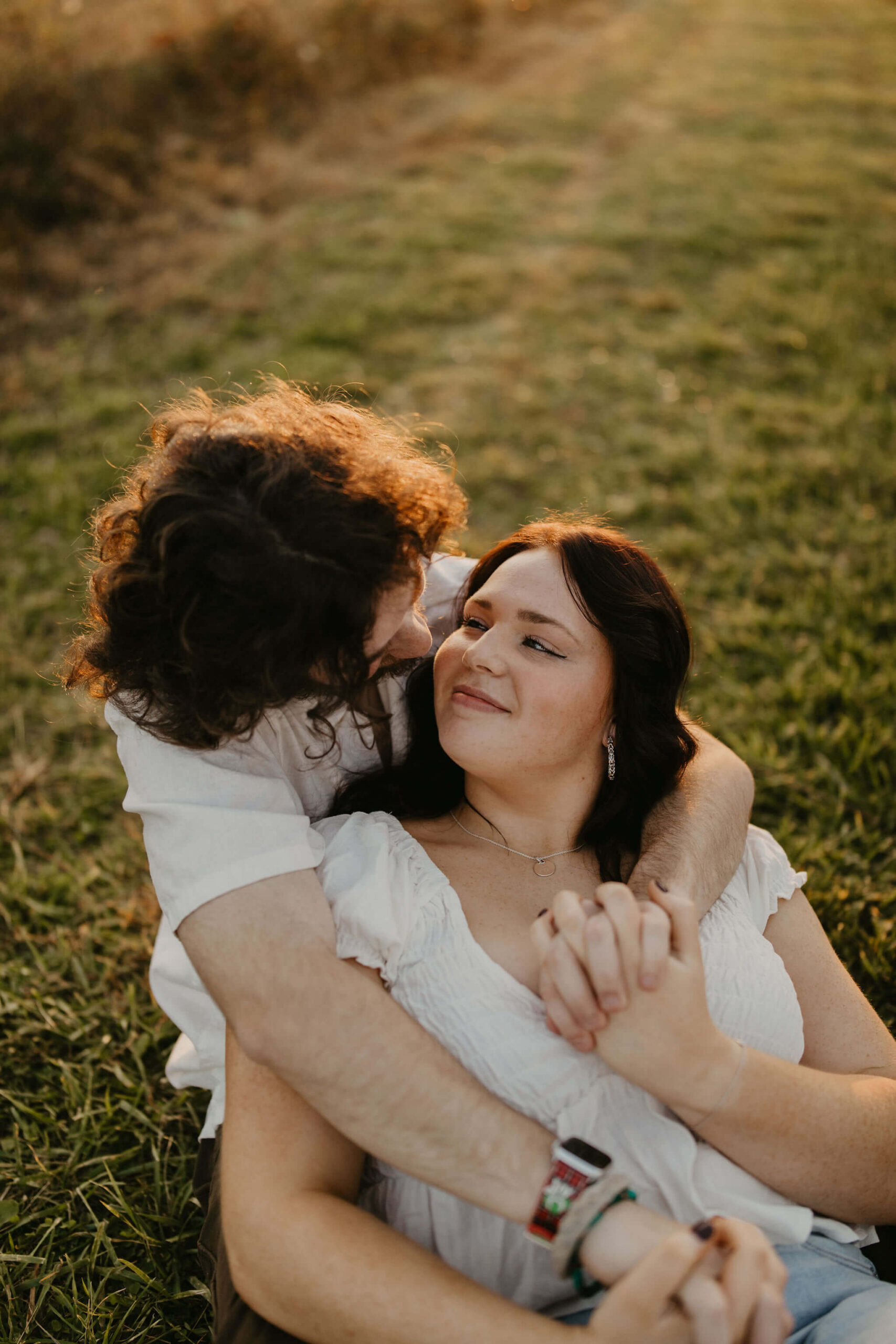 a couple sitting in a field cuddling. his arms are wrapped around her and they are smiling at each other