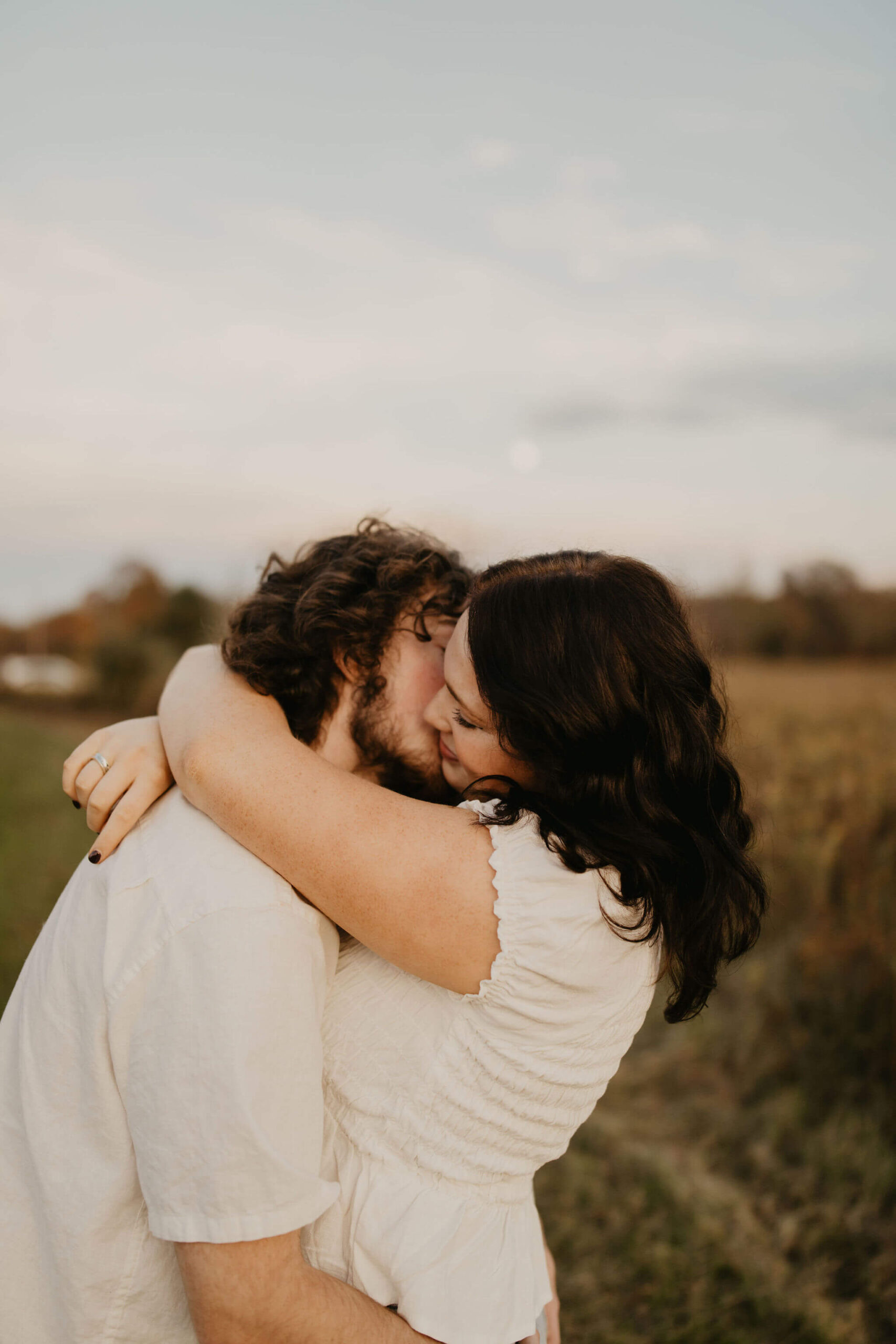 a boy kissing his girlfriend on the cheek in a field at sunset. the moon is softly rising behind them.