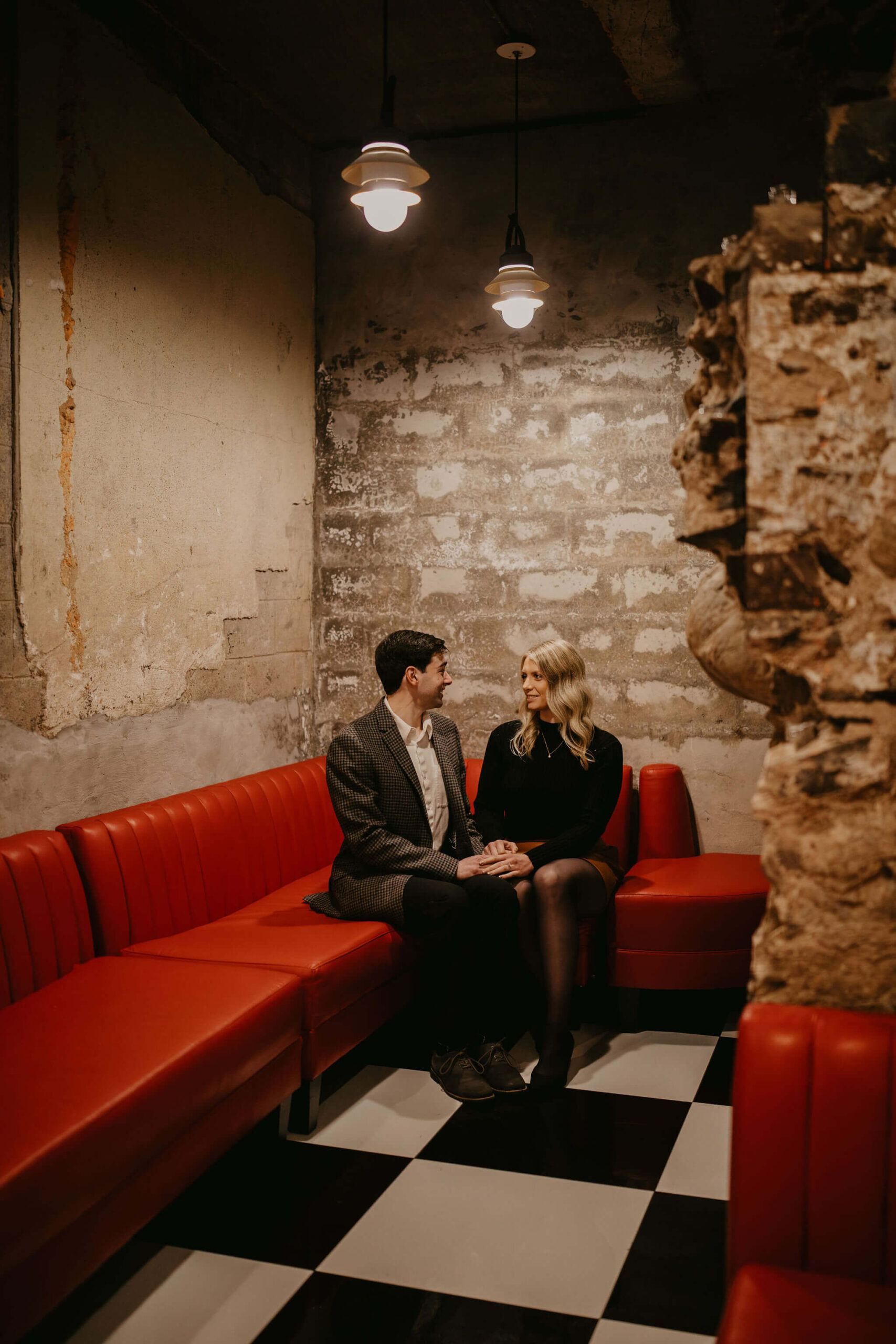 man (brown hair, brown checkered suit) and woman (long blonde hair, black shirt and brown skirt) sitting on a red leather couch holding hands and smiling at each other.