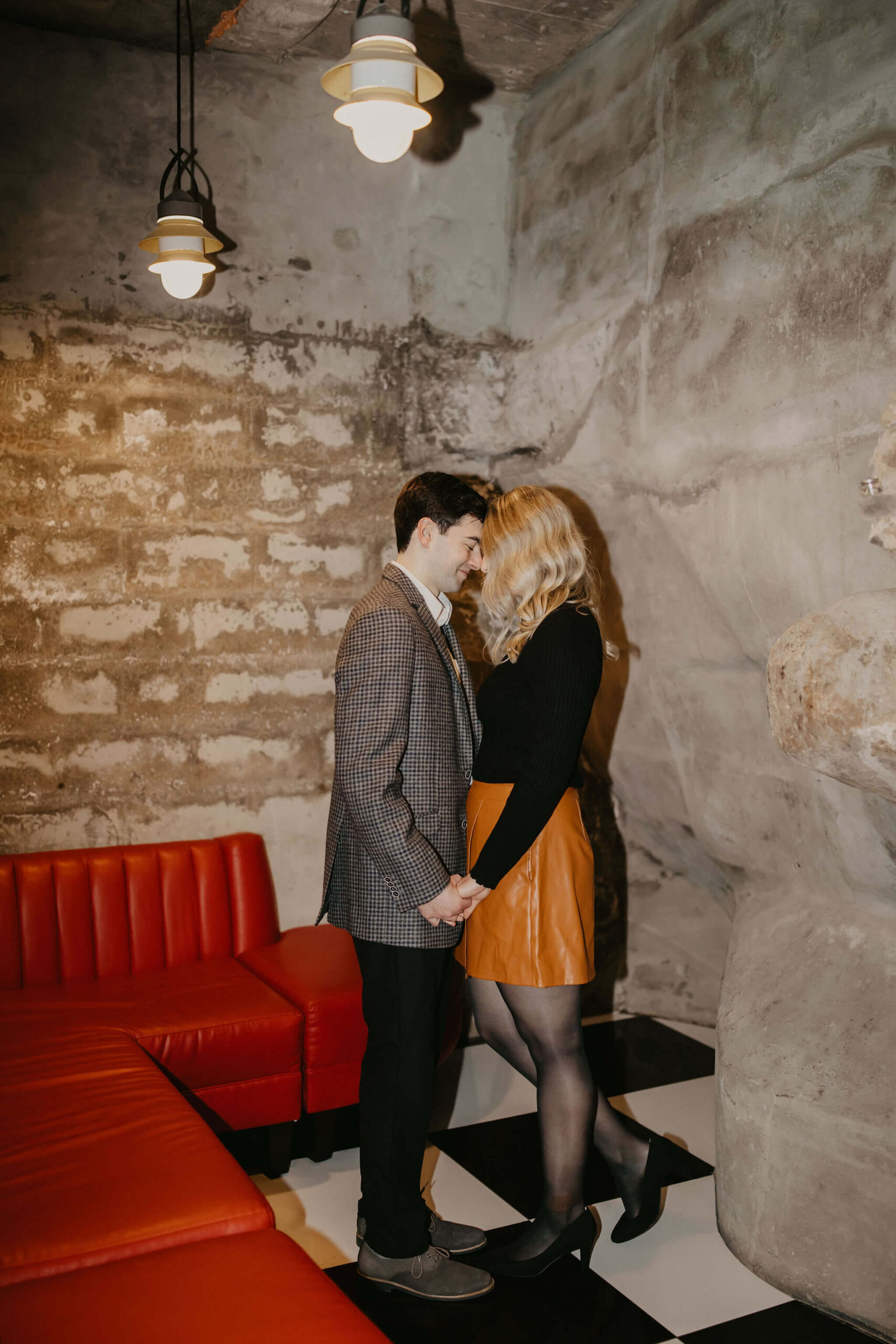 man (brown hair, brown checkered suit) and woman (long blonde hair, black shirt and brown skirt) holding hands and leaning together with eyes closed