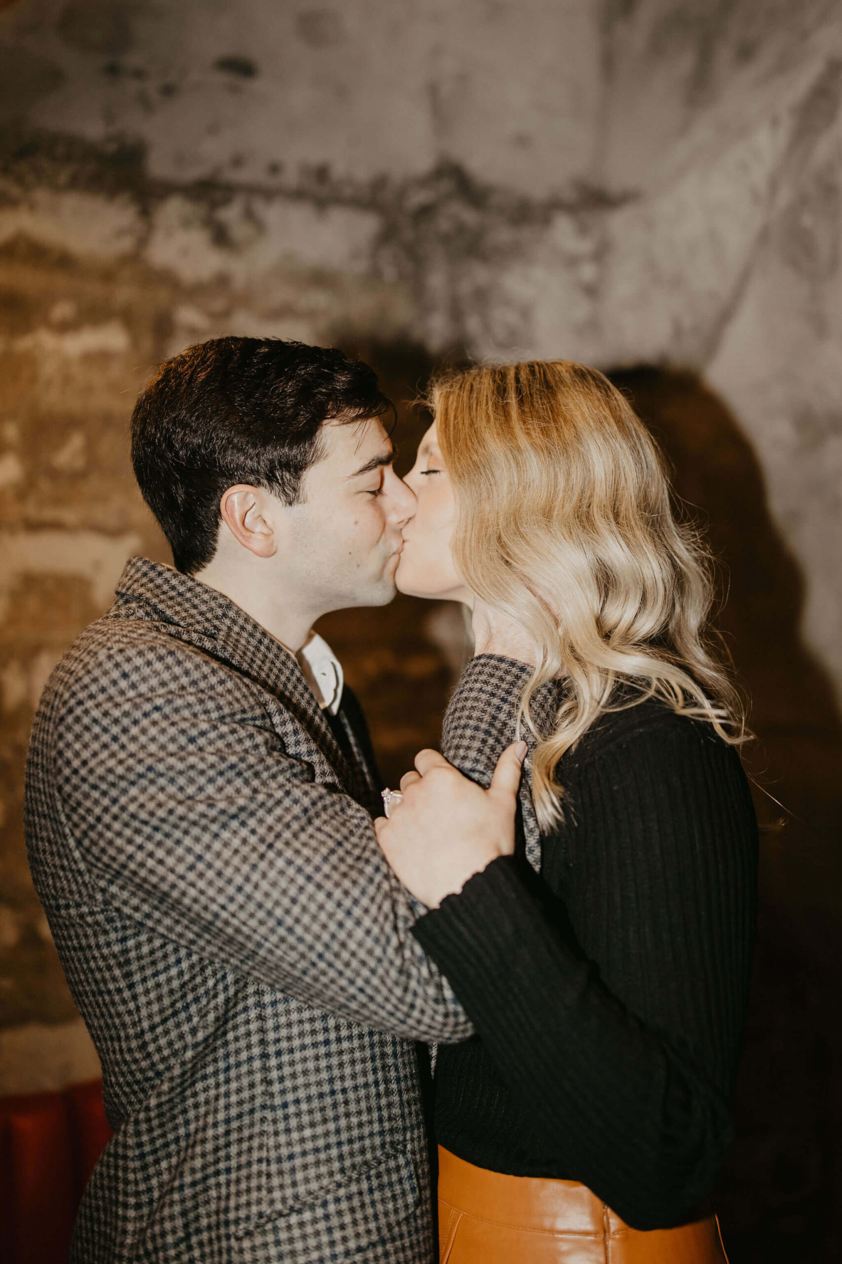 man (brown hair, brown checkered suit) holding woman woman's face (long blonde hair, black shirt and brown skirt) and kissing her. She holds his arm with her left hand to show her engagement ring.