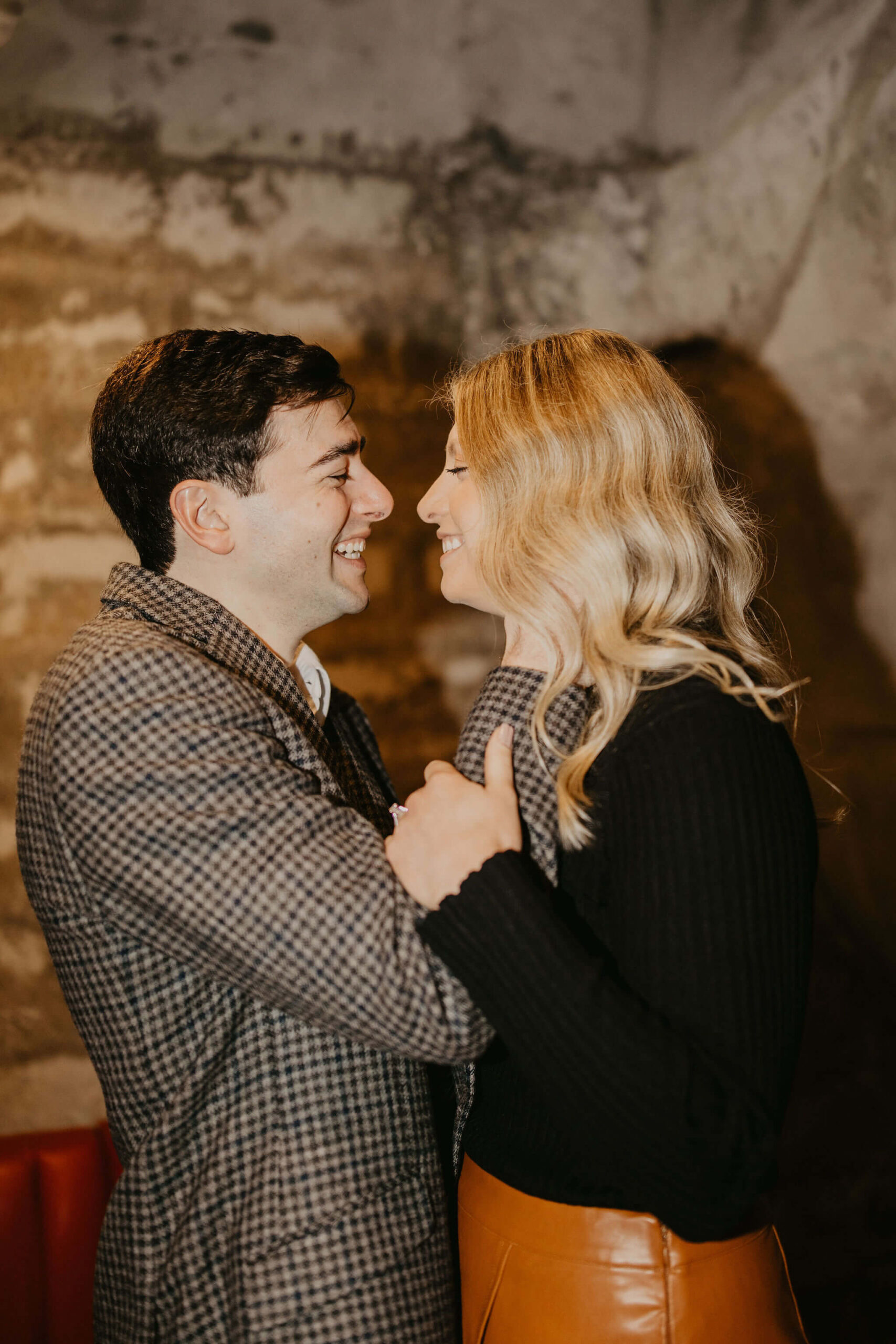 close up of a man (brown hair, brown checkered suit) and woman (long blonde hair, black shirt and brown skirt) smiling at each other and holding each other