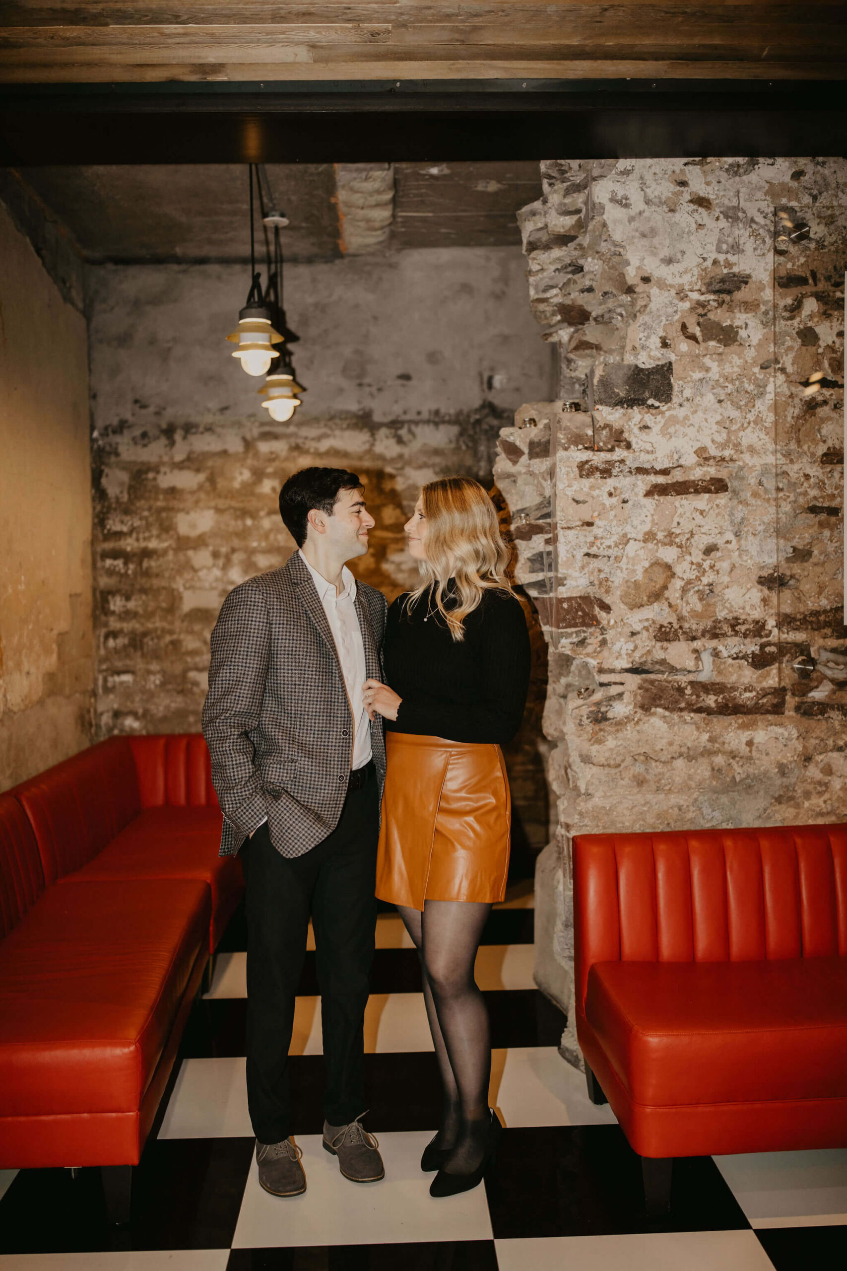 man (brown hair, brown checkered suit) and woman (long blonde hair, black shirt and brown skirt) in front of a red leather couch and stone wall, standing and looking at each other