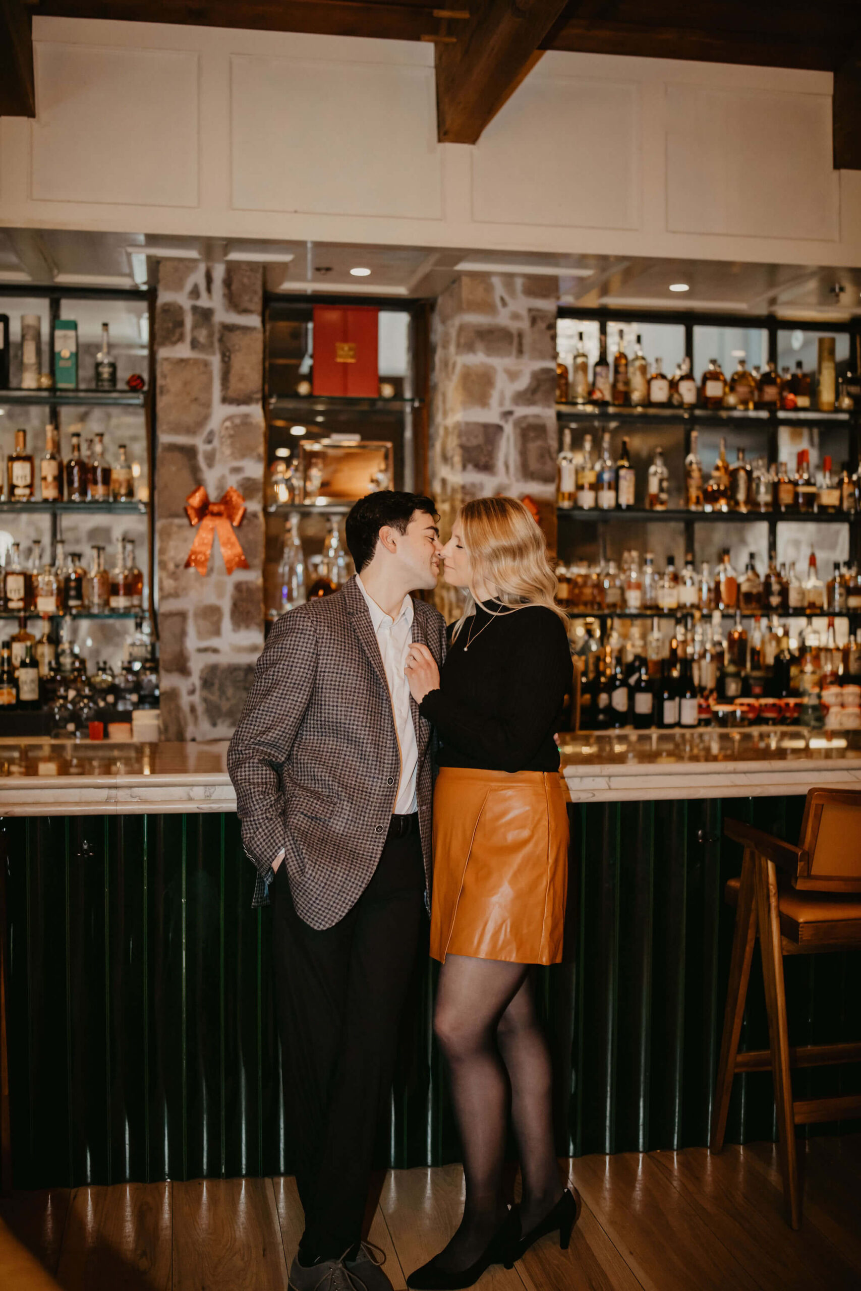 man and woman standing against a bar with a full liquor shelf behind it, she is holding the lapel of his suit and bringing him in for a kiss