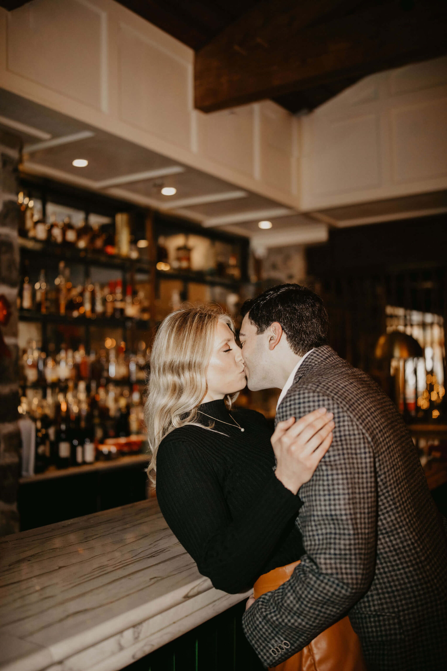 close up image of a man kissing a woman with her back pressed against a bar, a full liquor shelf visible behind them