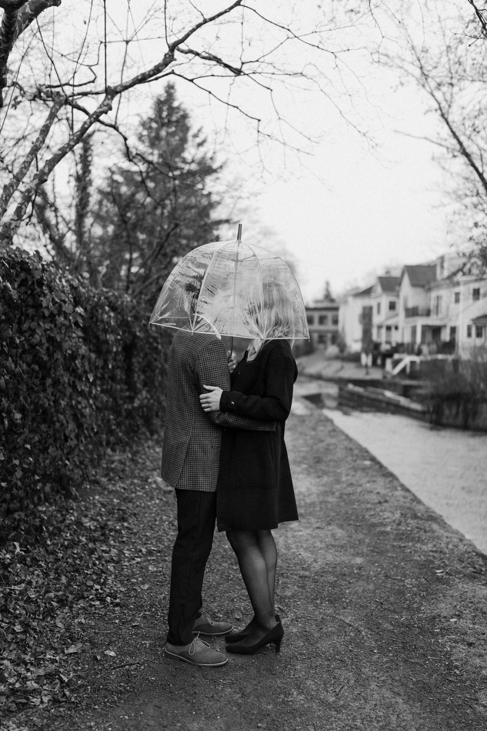 black and white image of a man and woman holding each other, with a clear umbrella held over their heads and obscuring the view of their faces