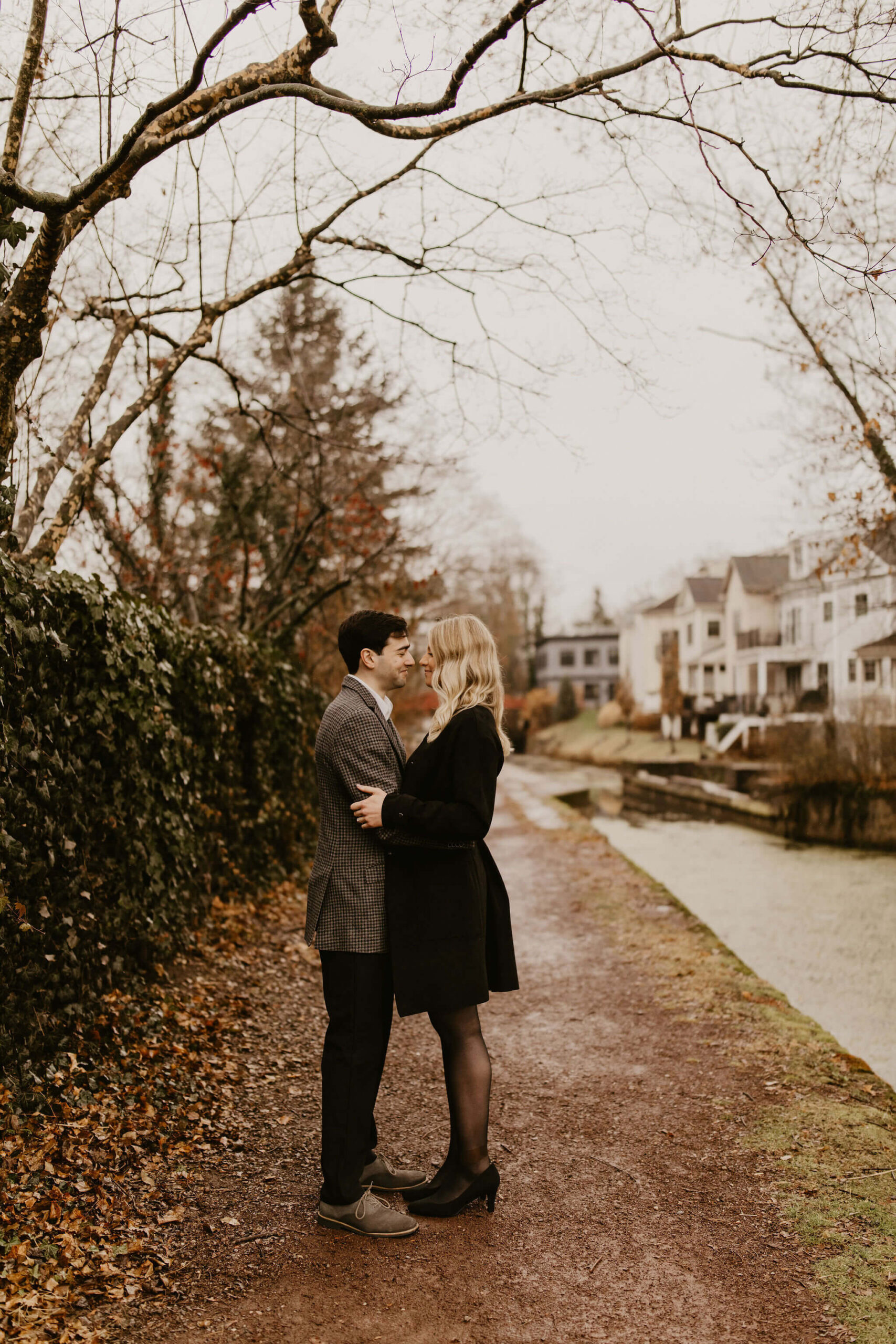 man (brown hair, brown checkered suit) and woman (long blonde hair, long black jacket and tights) embracing next to a river and smiling at each other