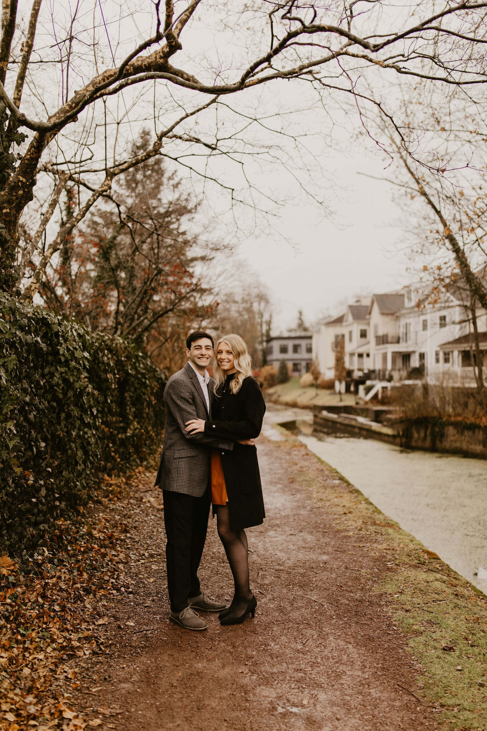 man (brown hair, brown checkered suit) and woman (long blonde hair, black shirt and brown skirt) holding each other on a walking trail next to a river, looking at the camera and smiling