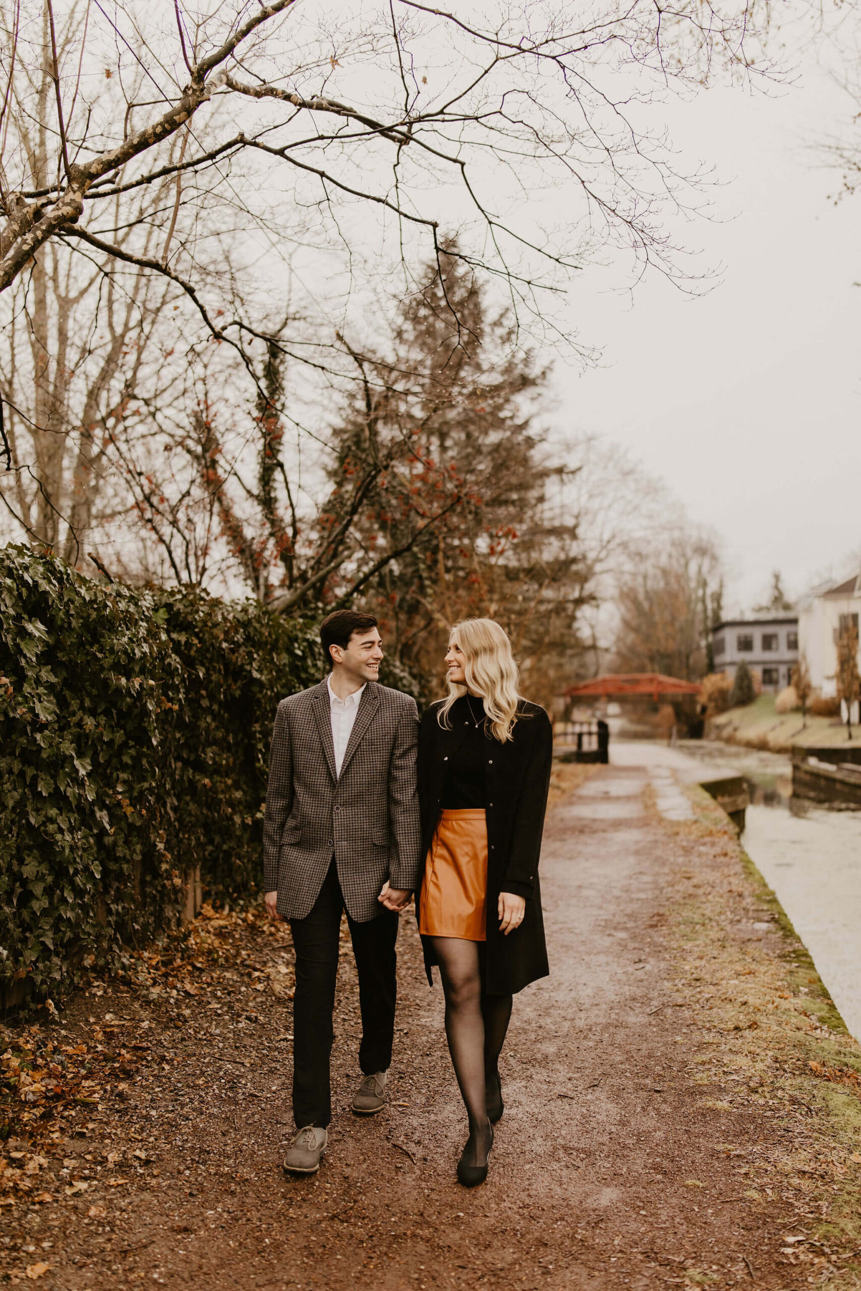 man (brown hair, brown checkered suit) and woman (long blonde hair, black shirt and brown skirt) walking toward the camera along a path and smiling at each other