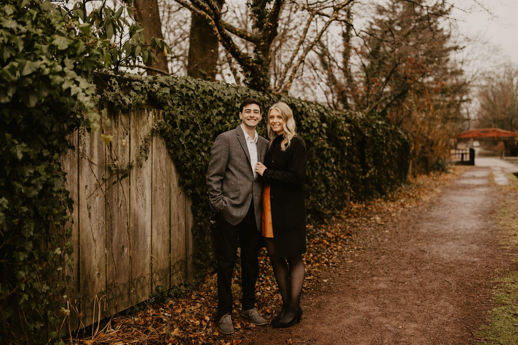 man (brown hair, brown checkered suit) and woman (long blonde hair, black shirt and brown skirt) holding each other in front of a fence with ivy growing on it, looking at the camera and smiling