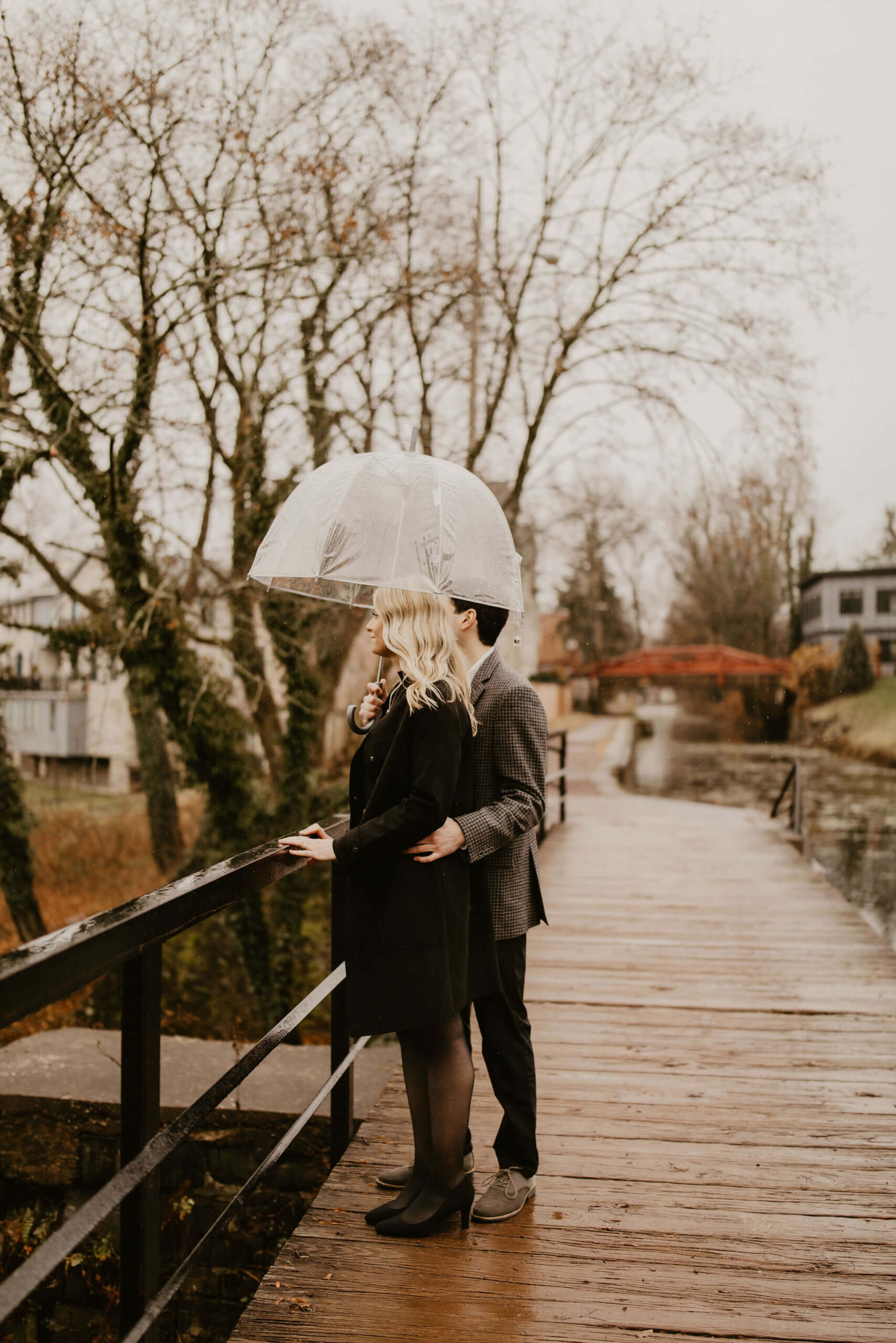 man with his arm around his fiance's back, looking off camera to the left while standing on a bridge, holding a clear umbrella over them both