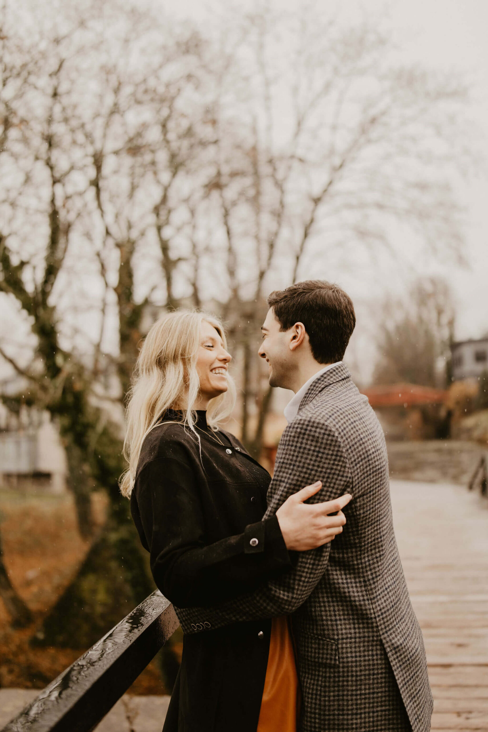 a couple embracing on a bridge, with her back pressed against the railing. They are looking at each other and smiling.