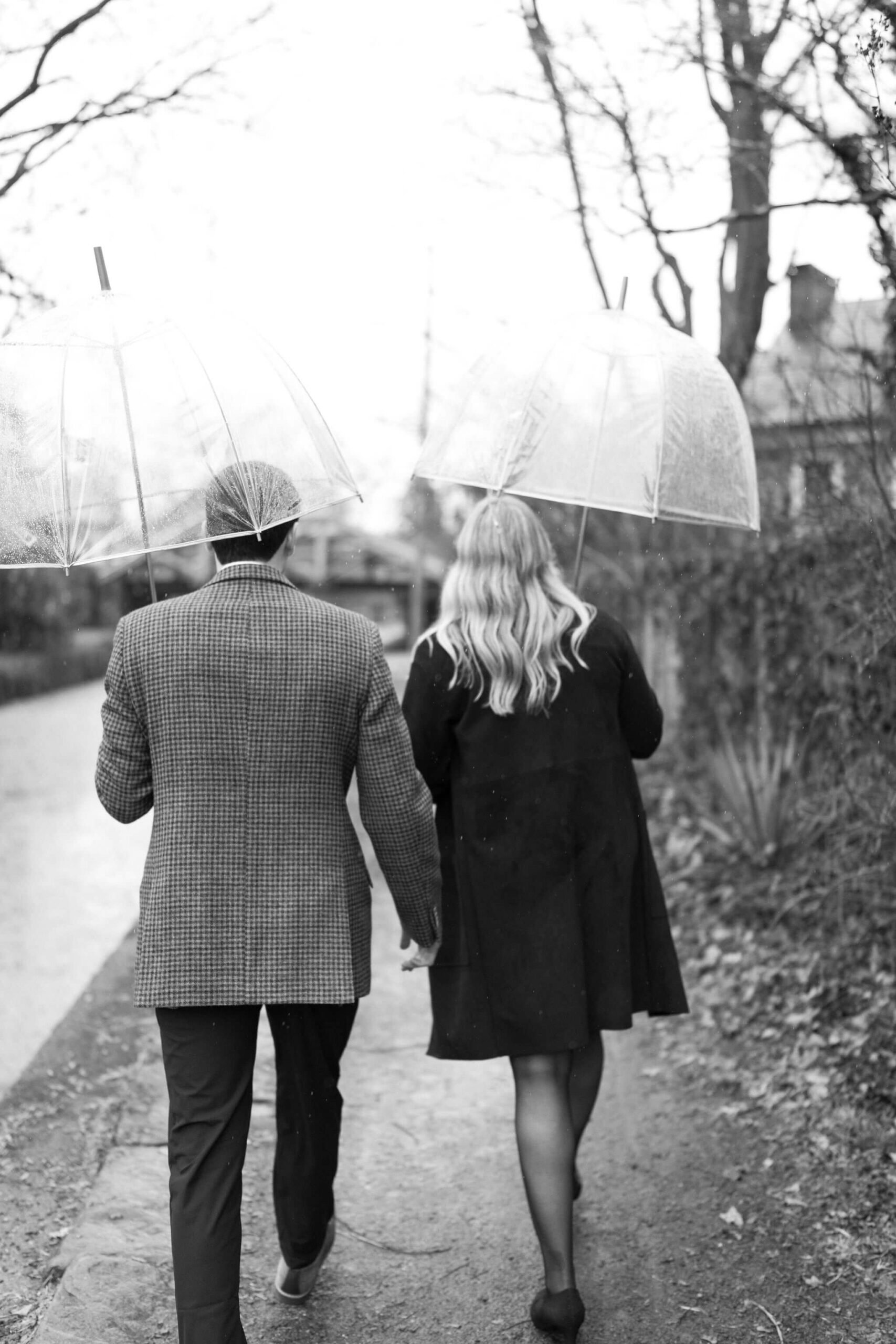 black and white image of a couple walking away from the camera, both wearing winter jackets and holding clear umbrellas over their head as raindrops fall around them