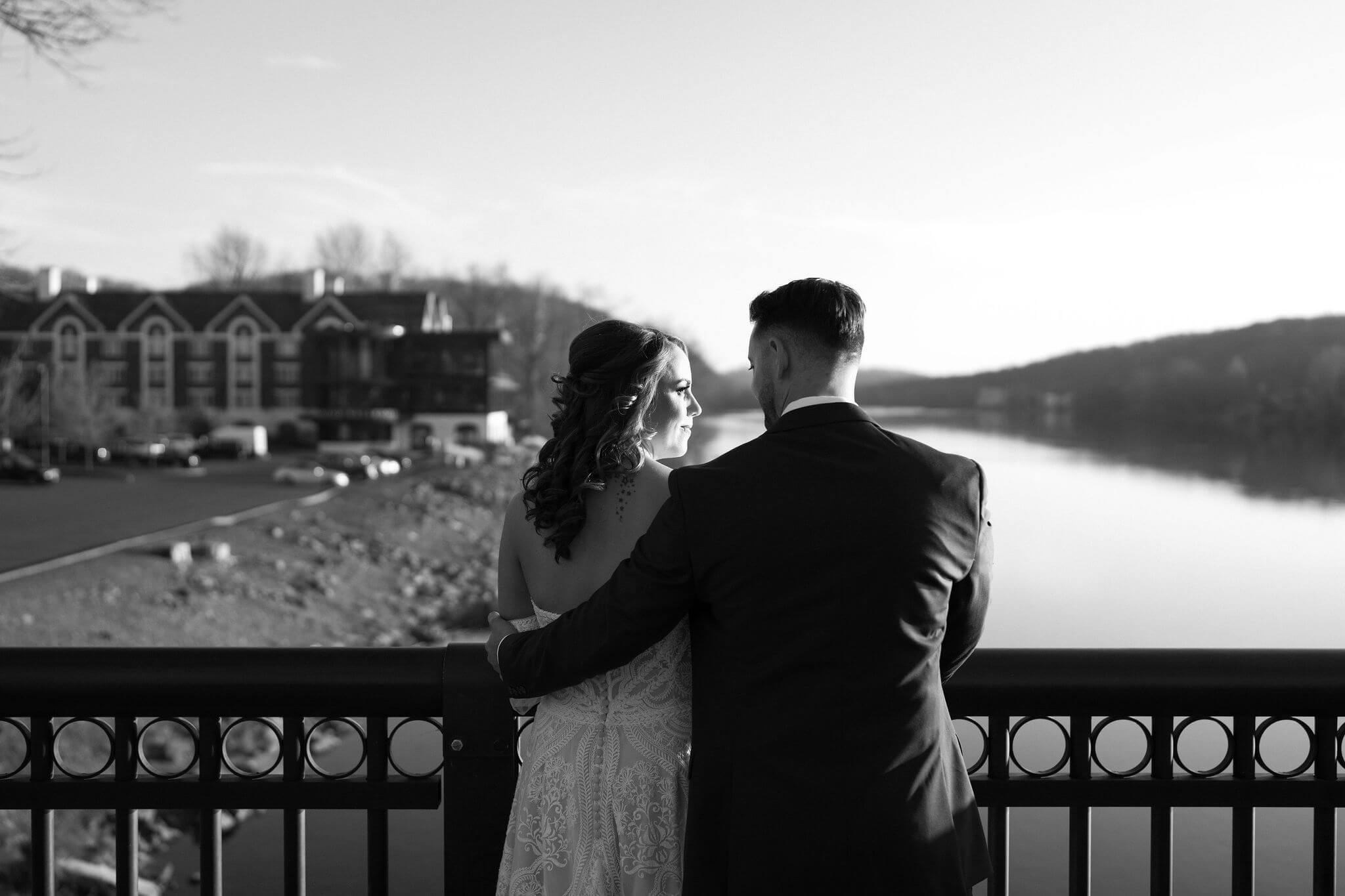 black and white image of a bride and groom looking at each other while standing against the railing of a bridge overlooking water