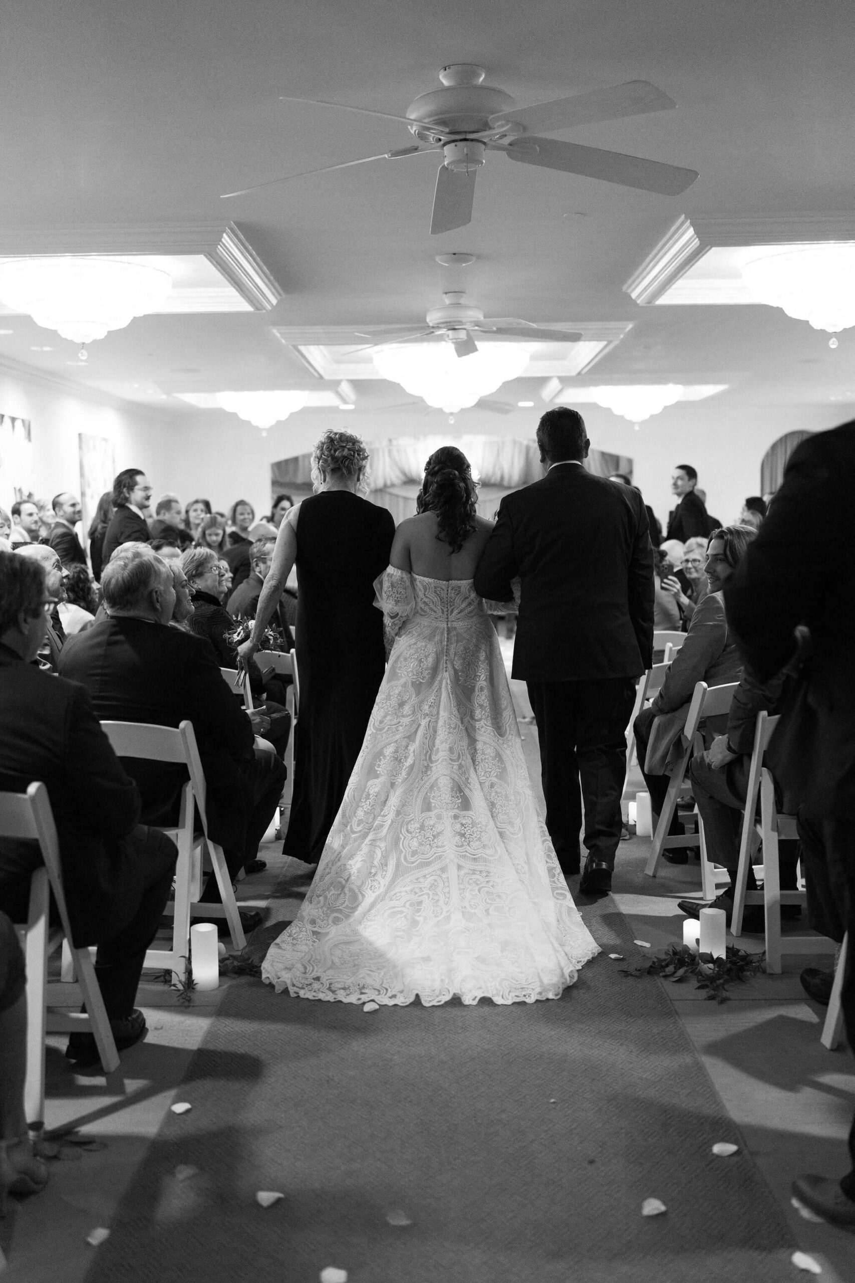 black and white image of a bride at her wedding being walked down the aisle by her mom and dad, on either side of her