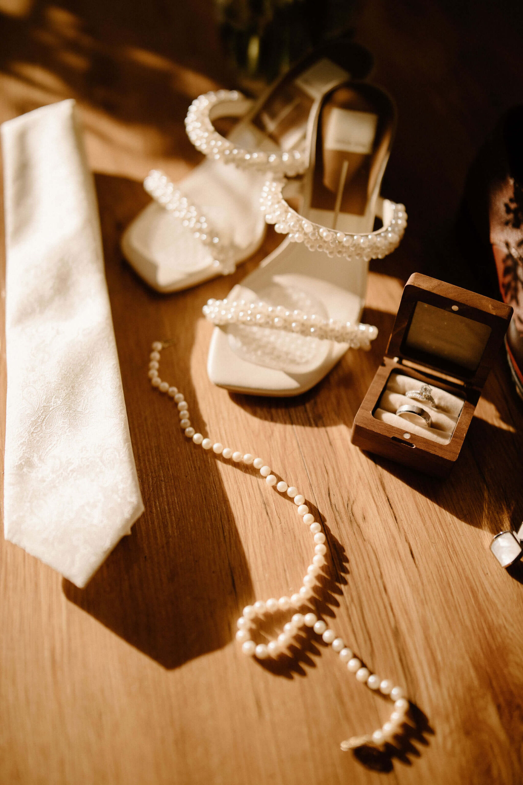 close up detail photo of pearl-studded heels, a cream colored tie, pearl necklace, and an engagement ring, diamond-studded wedding band, and silver wedding band in a wooden box