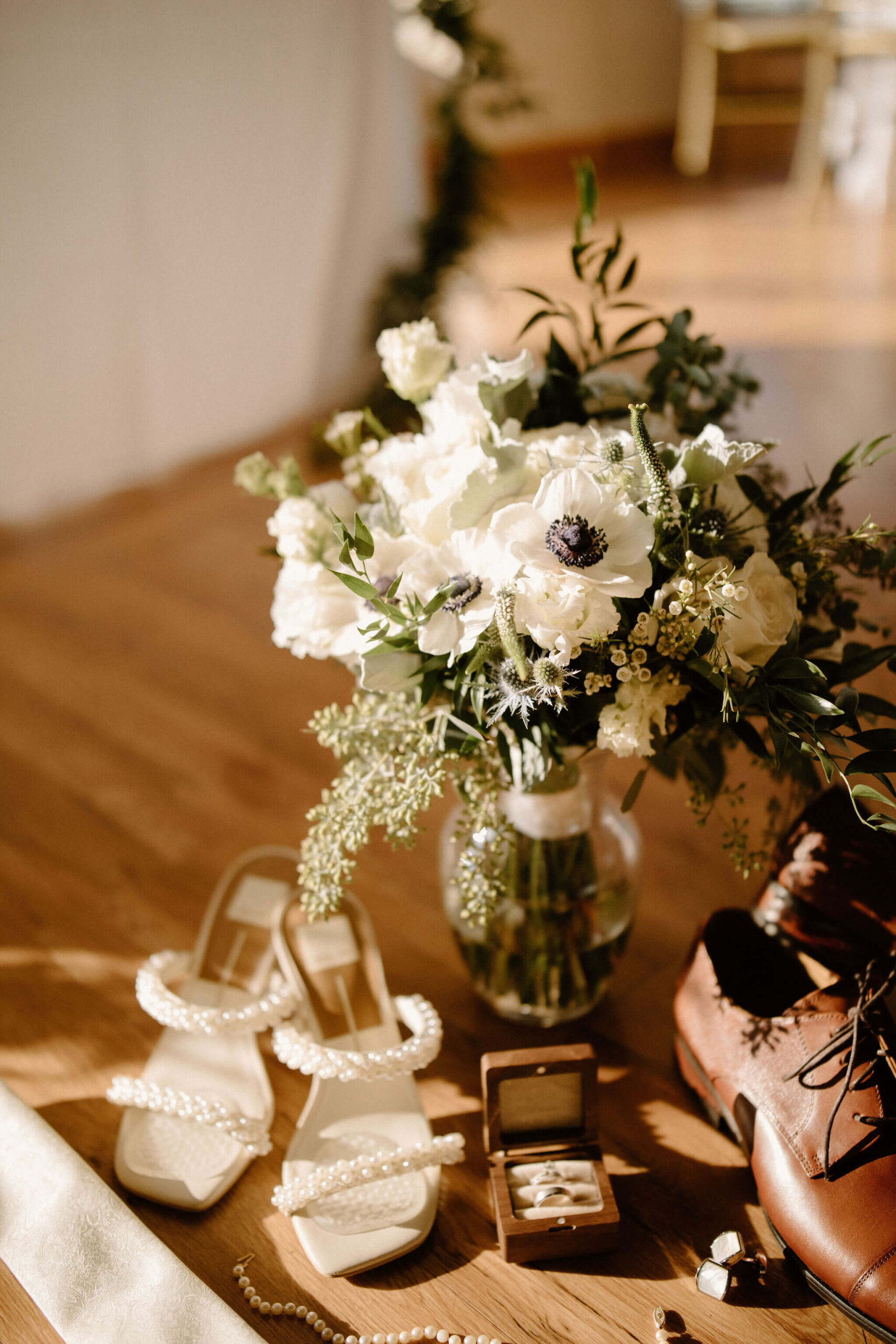 bridal bouquet in a vase with white poppies and greenery, surrounded by wedding rings in a wooden box, brown leather dress shoes, and pearl studded white heels.