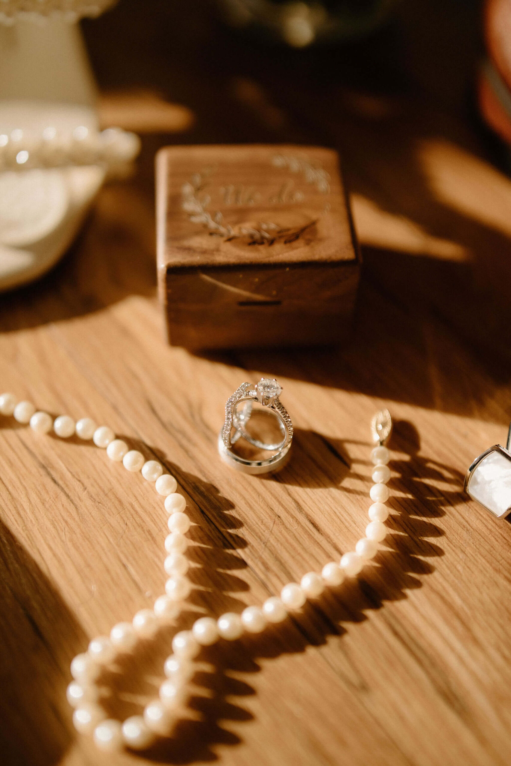 a close up of a diamond engagement ring and diamond-studded wedding band, balanced on a silver wedding band, surrounded by a pearl necklace and wooden ring box