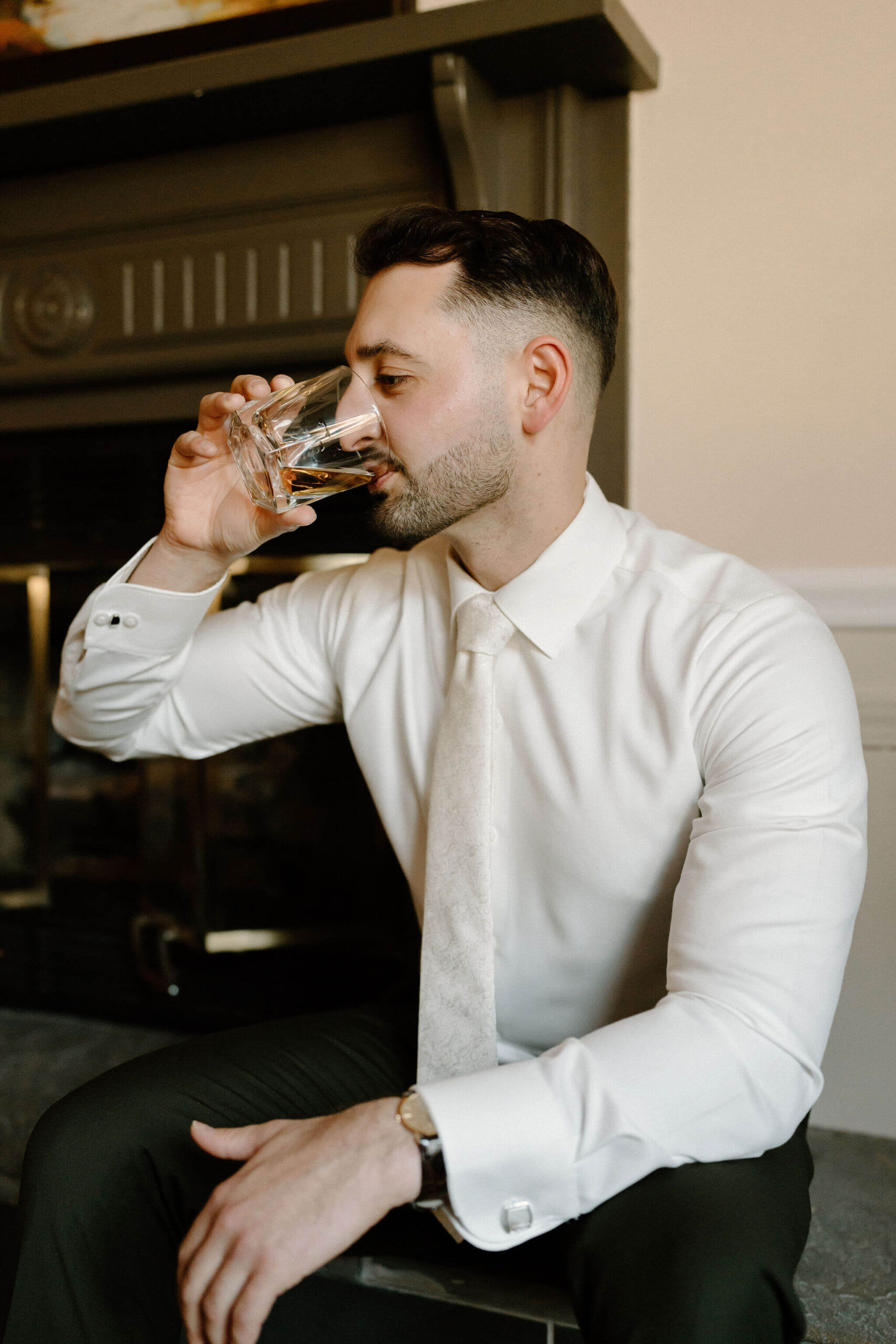 groom (brown hair, white button down shirt, and ivory tie) sitting and sipping a brown liquor