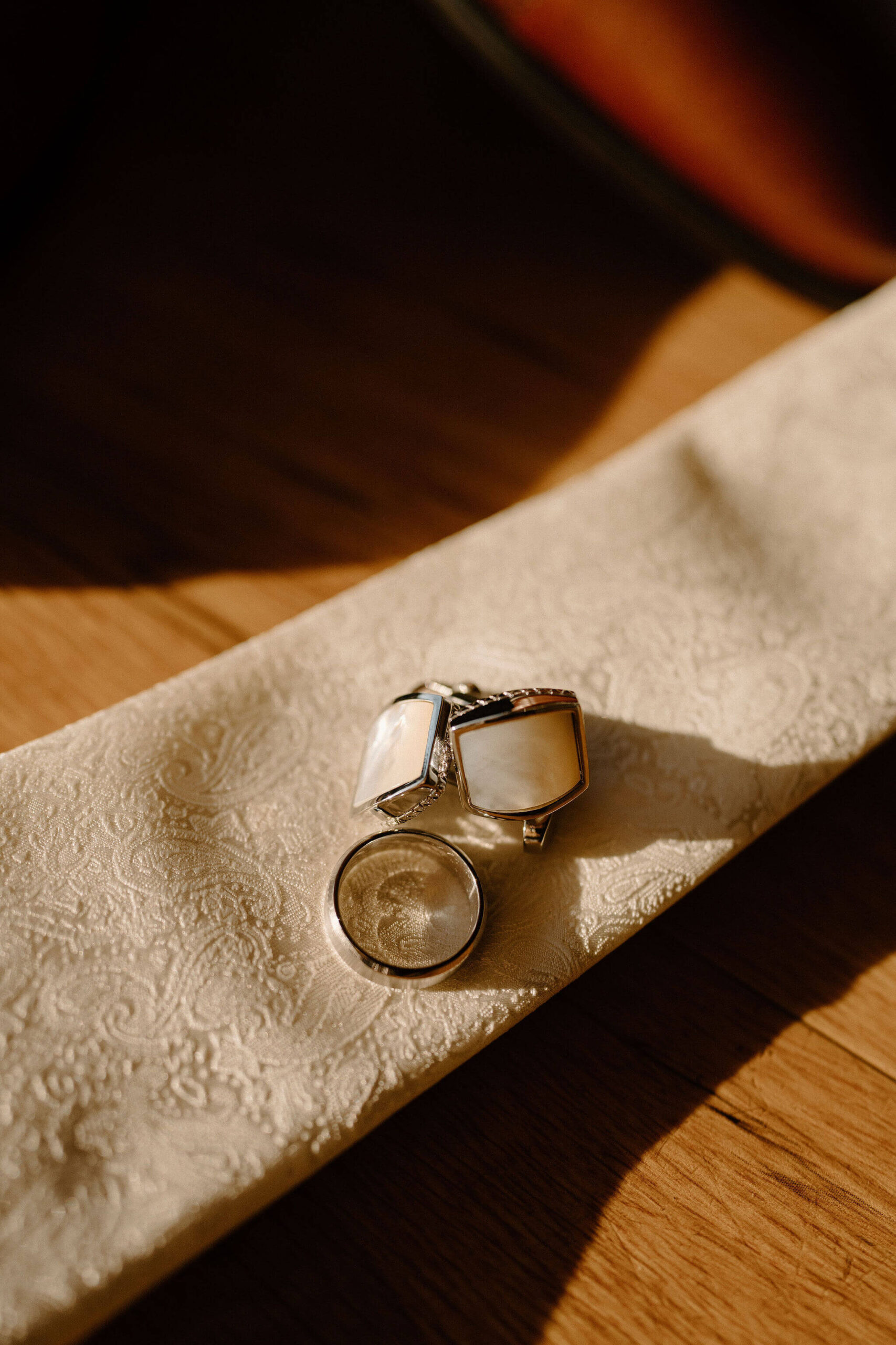 Close up image of ivory cuff links and a men's silver wedding band, arranged on a cream colored paisley tie