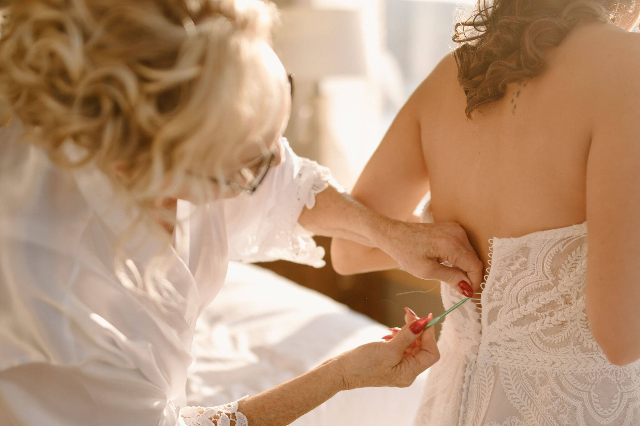 the mother of the bride in a white robe helping her daughter (the bride) button her wedding dress with a crochet hook