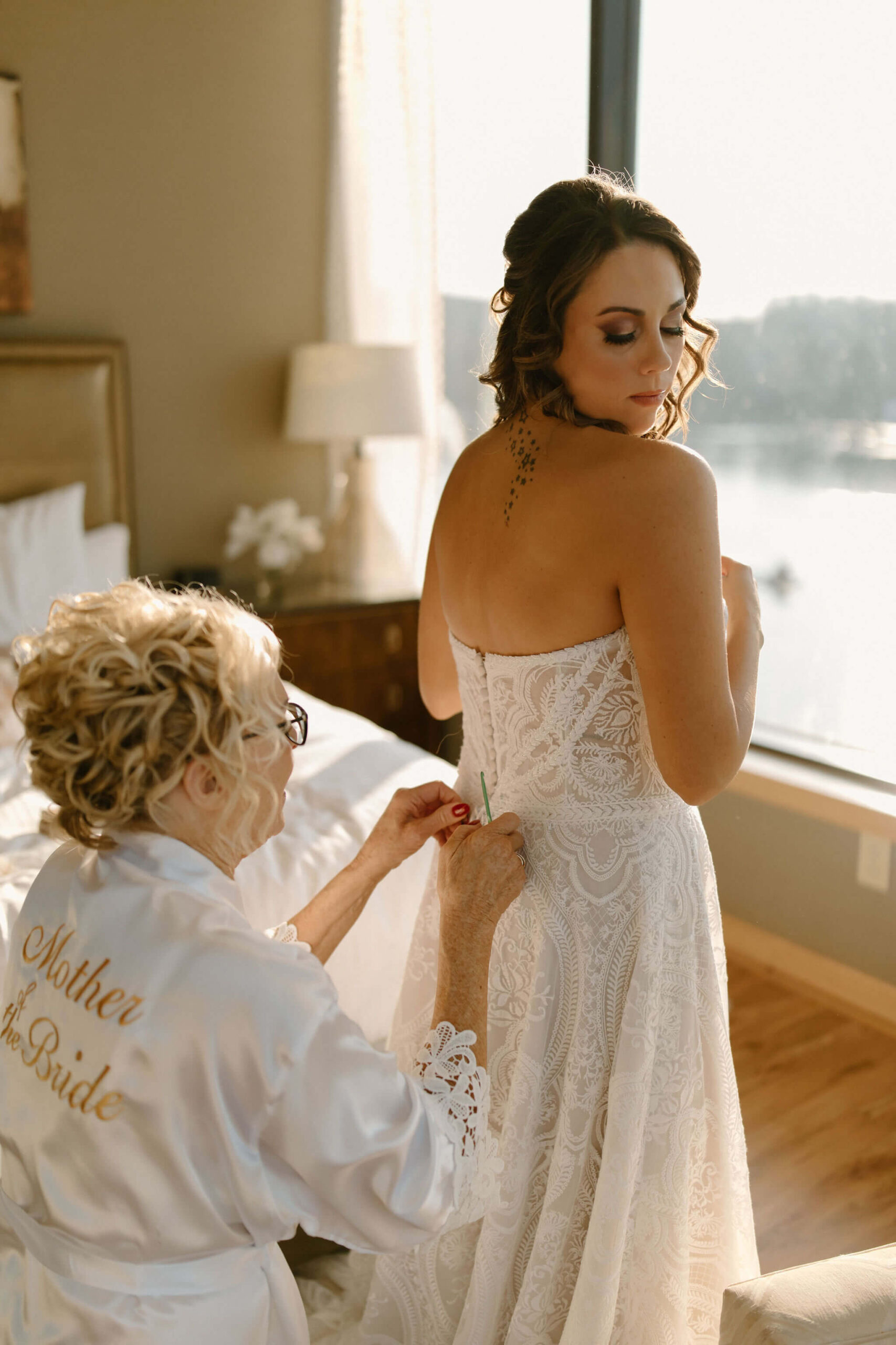 the mother of the bride in a white robe helping her daughter (the bride) button her wedding dress with a crochet hook