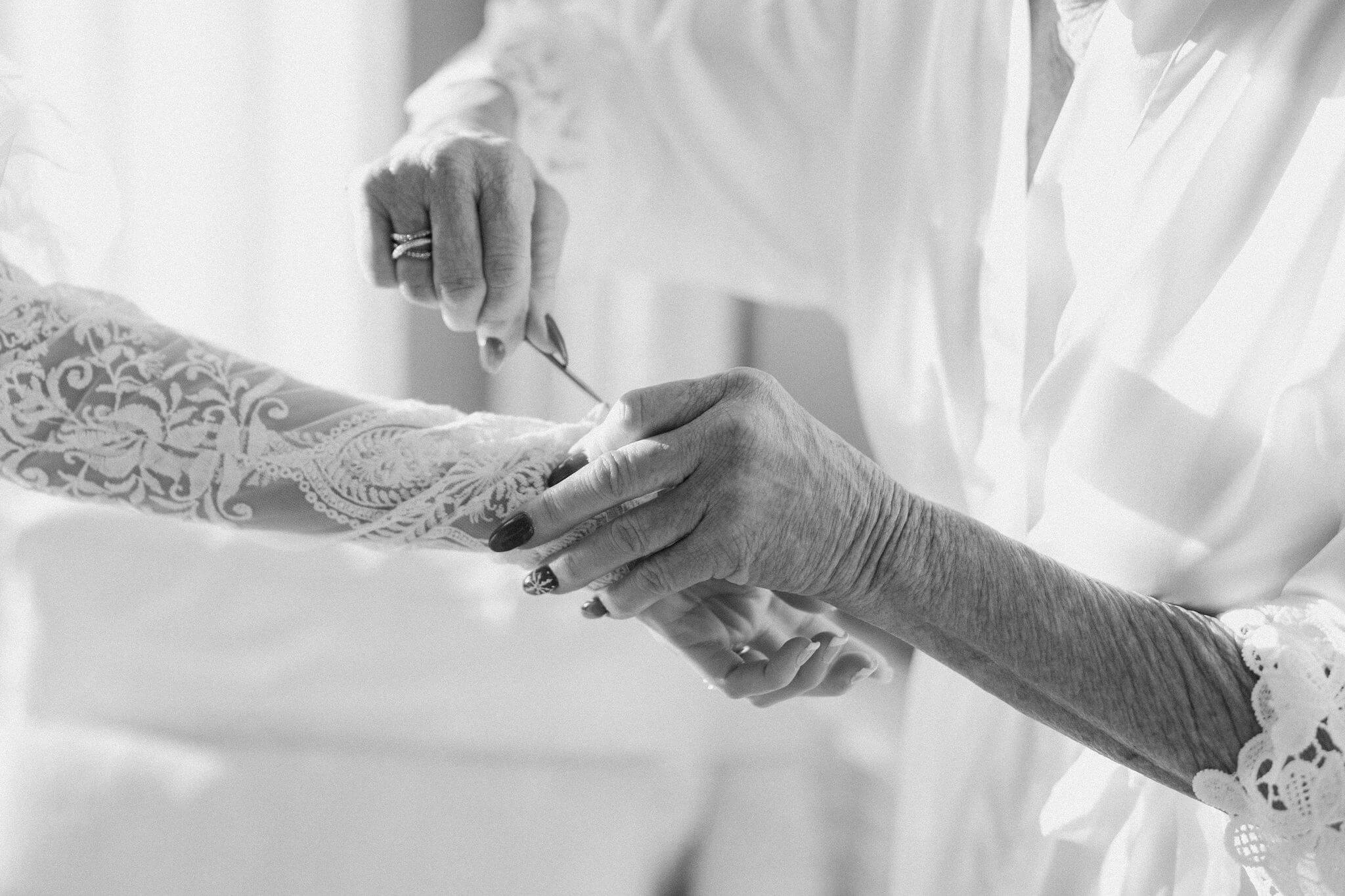 the mother of the bride in a white robe helping her daughter (the bride) button her wedding dress with a crochet hook