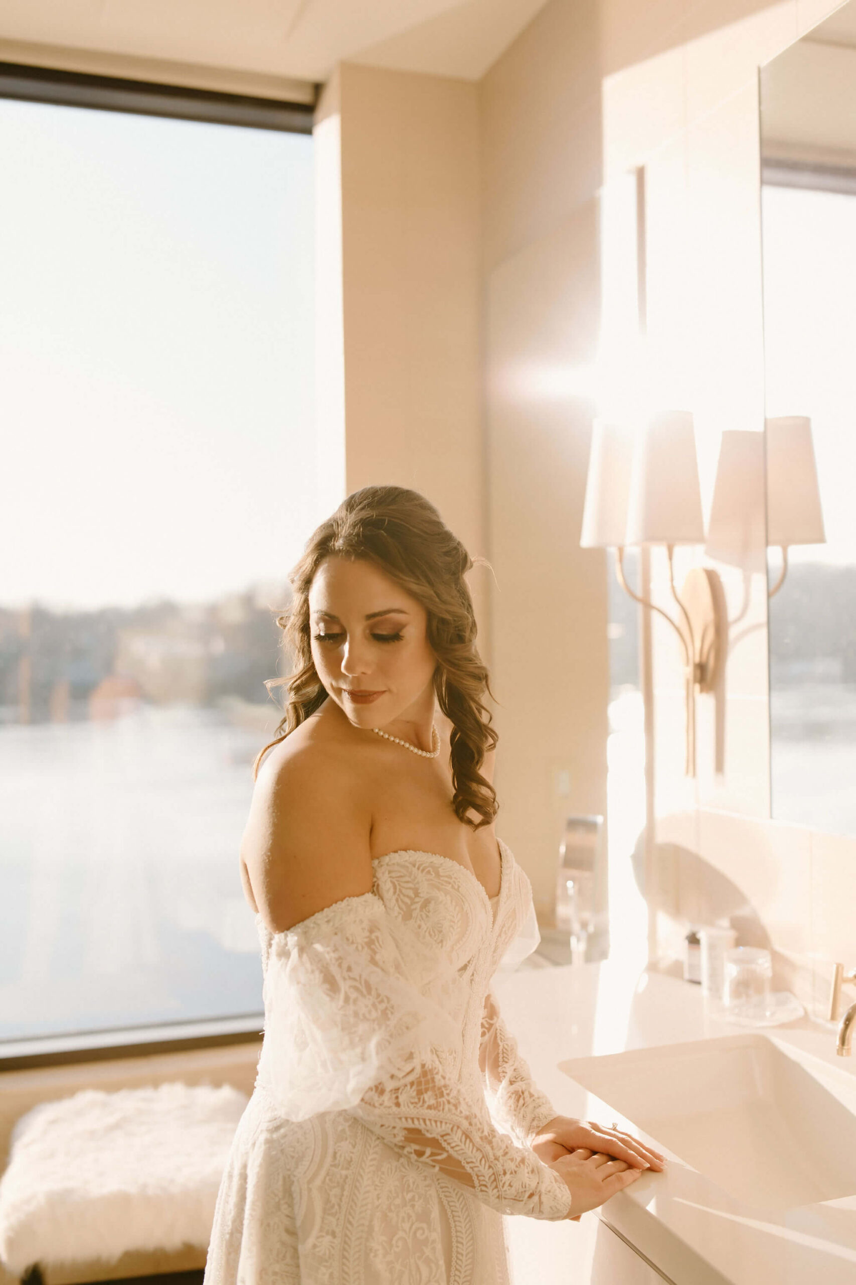 the bride in a bathroom, backlit by the setting sun, looking over her shoulder toward the camera with her eyes closed