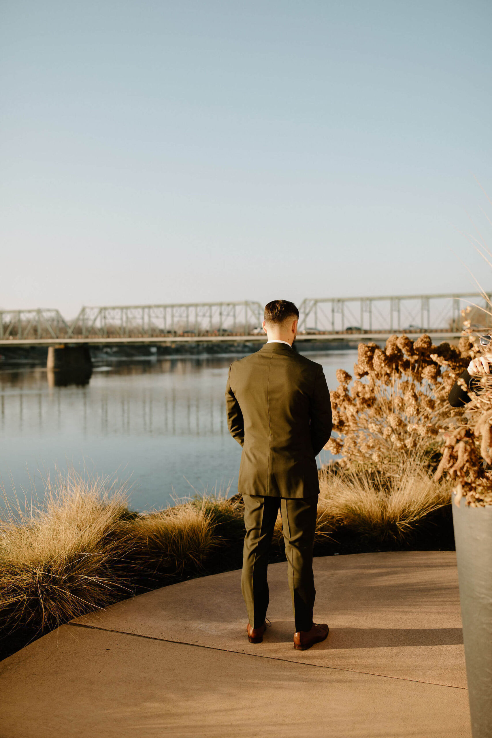 male groom (brown hair, green suit) facing away from the camera toward a walking bridge, waiting for the first look with his bride