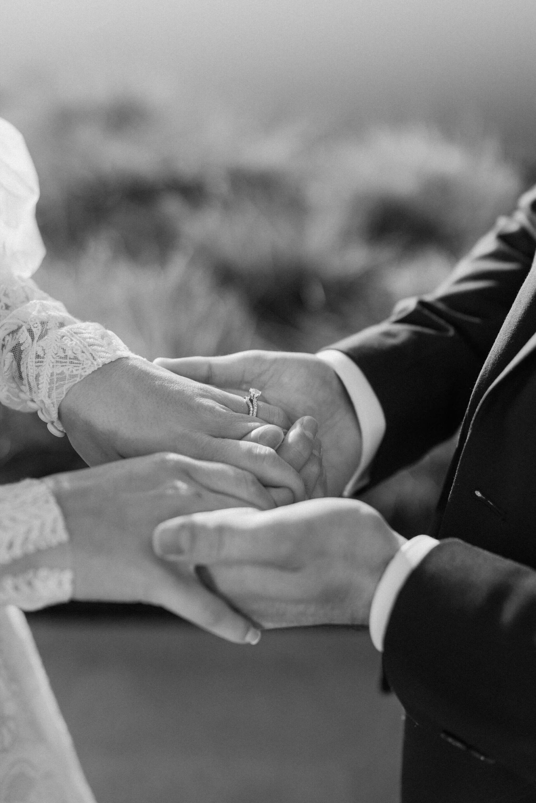 close up black and white image of a bride and groom holding hands, focused on her engagement ring and wedding band