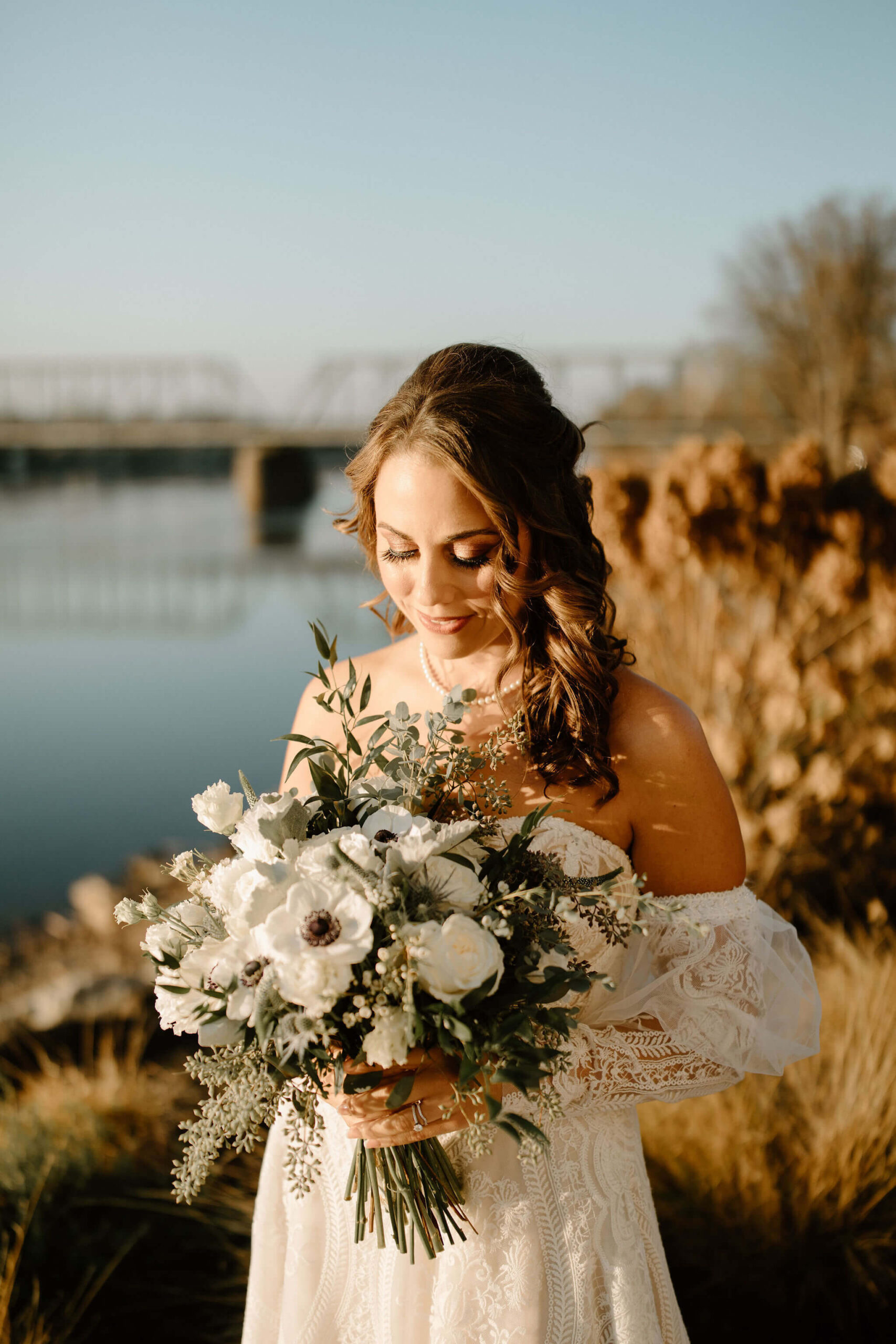 bride (brown hair, white dress) looking down at her bridal bouquet (white flowers with greenery)