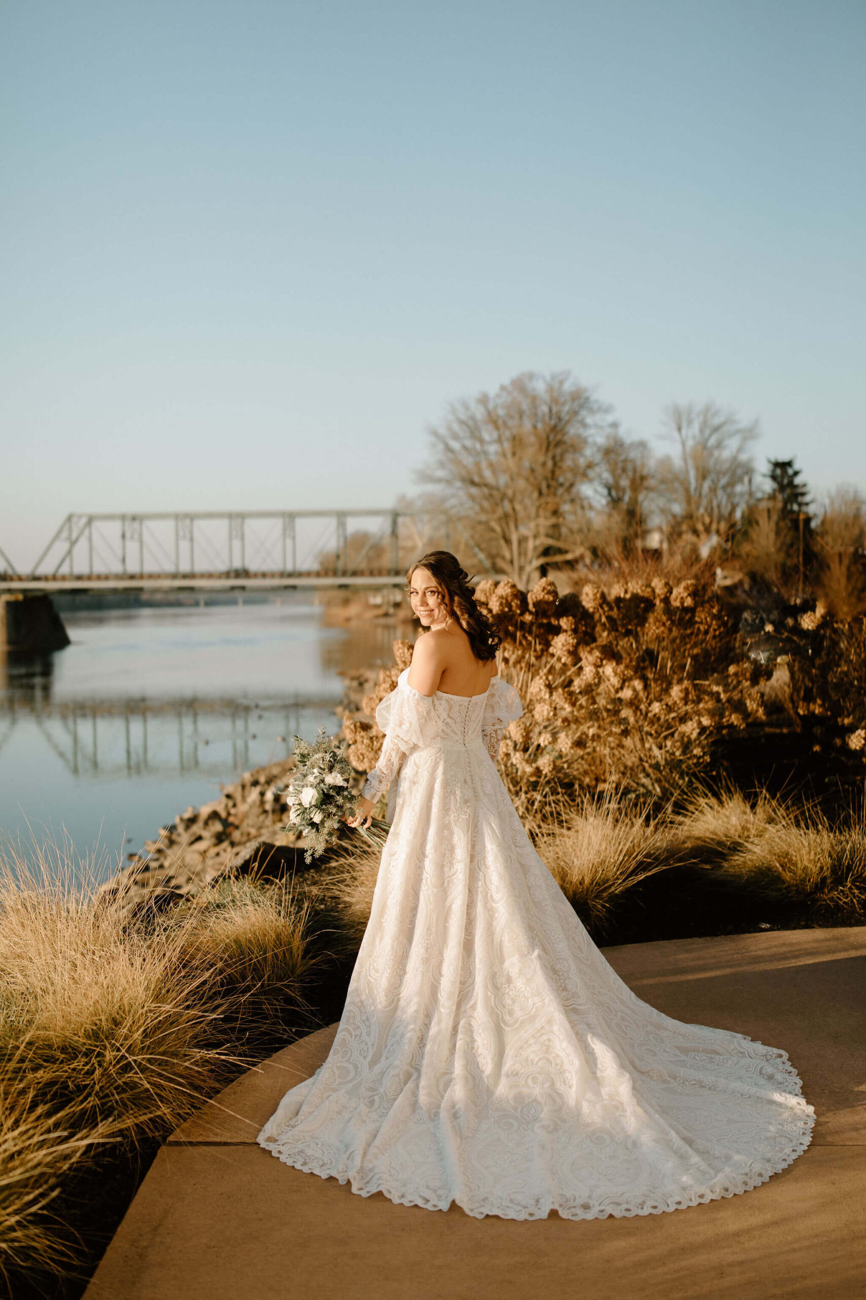 bride looking over her left shoulder toward the camera, off-white textured dress trailing behind her and a walking bridge over a river in the background