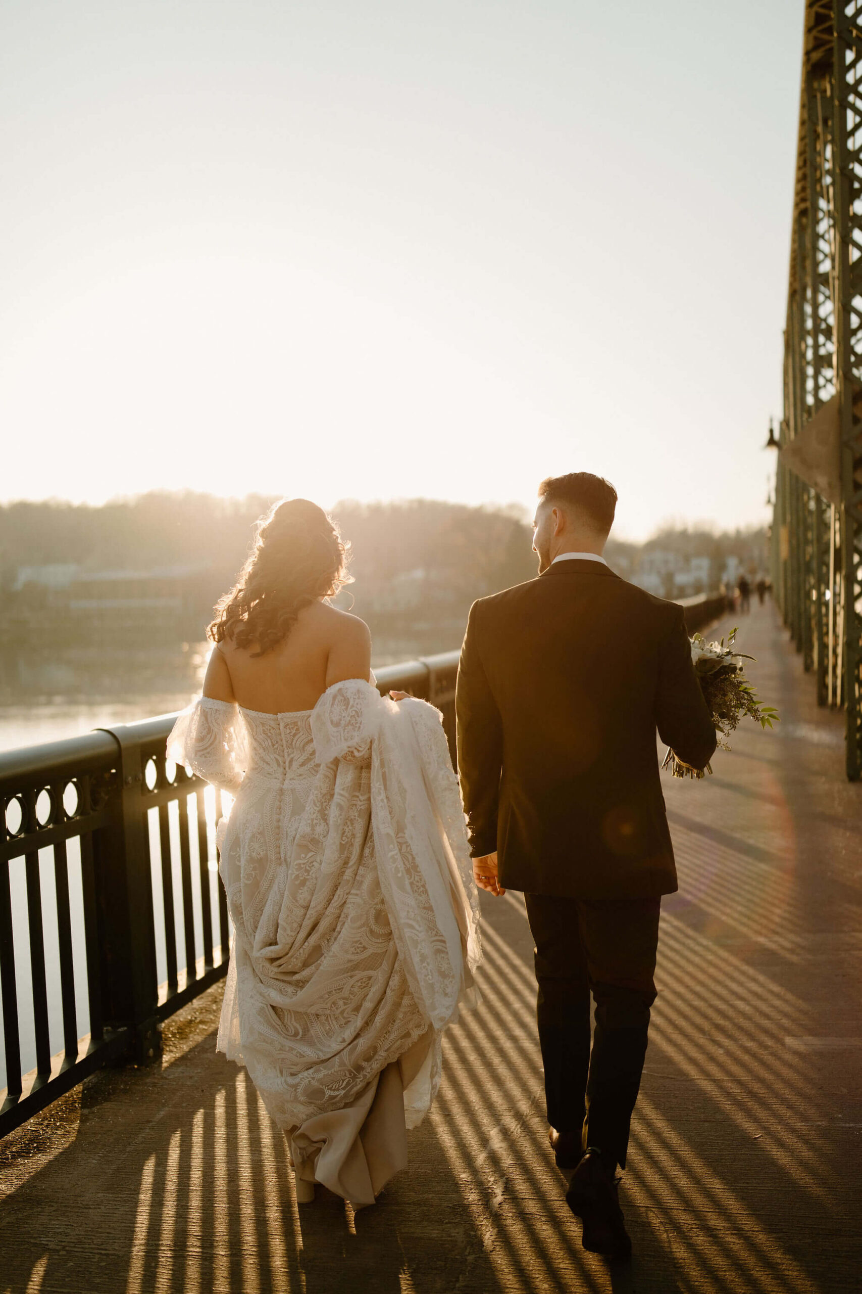 Bride (bohemian long sleeve white dress) and Groom (green suit) walking toward the sunset on a green bridge