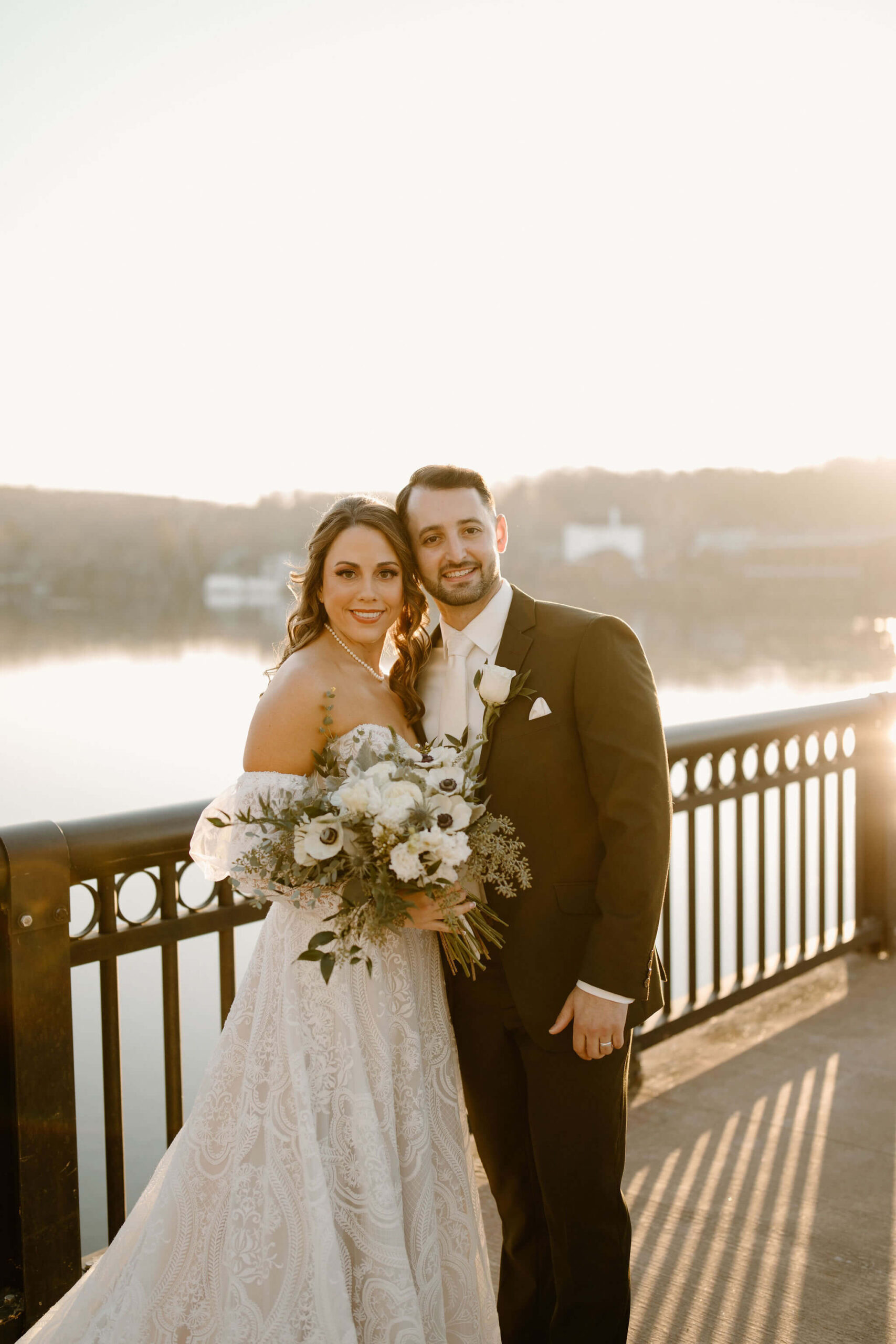 bride and groom smiling at the camera. She is holding flowers. They're standing on a bridge at sunset.