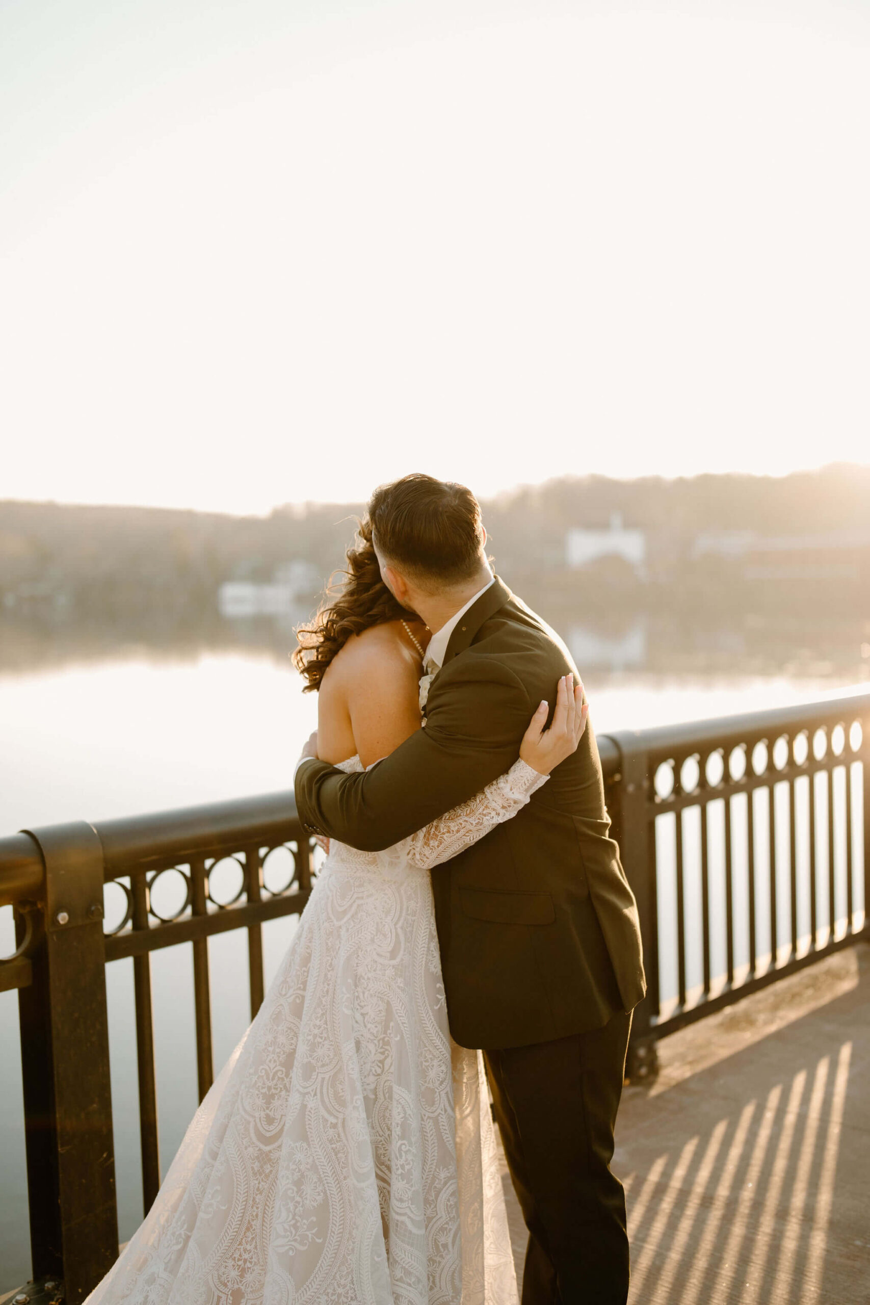 bride (boho white dress) and groom (green suit) embracing during sunset on a bridge overlooking a river