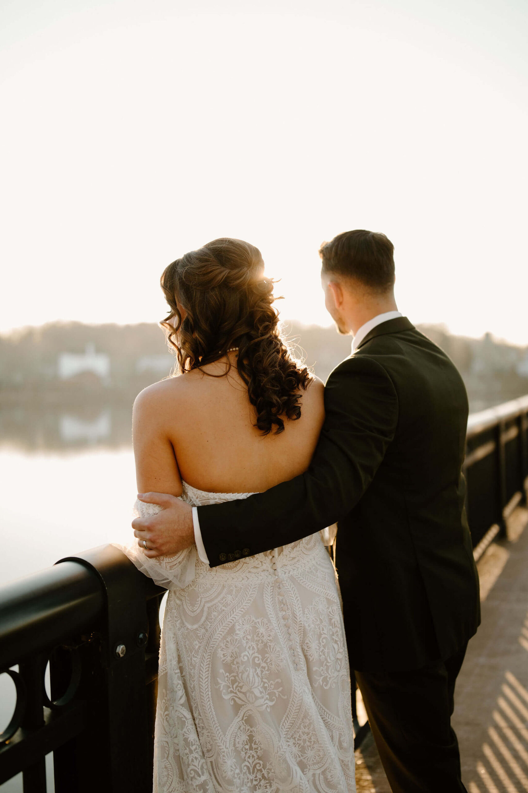 Groom with his arm wrapped around his bride, both facing away from the camera. They are standing against the railing of a bridge overlooking a river, watching the sunset.