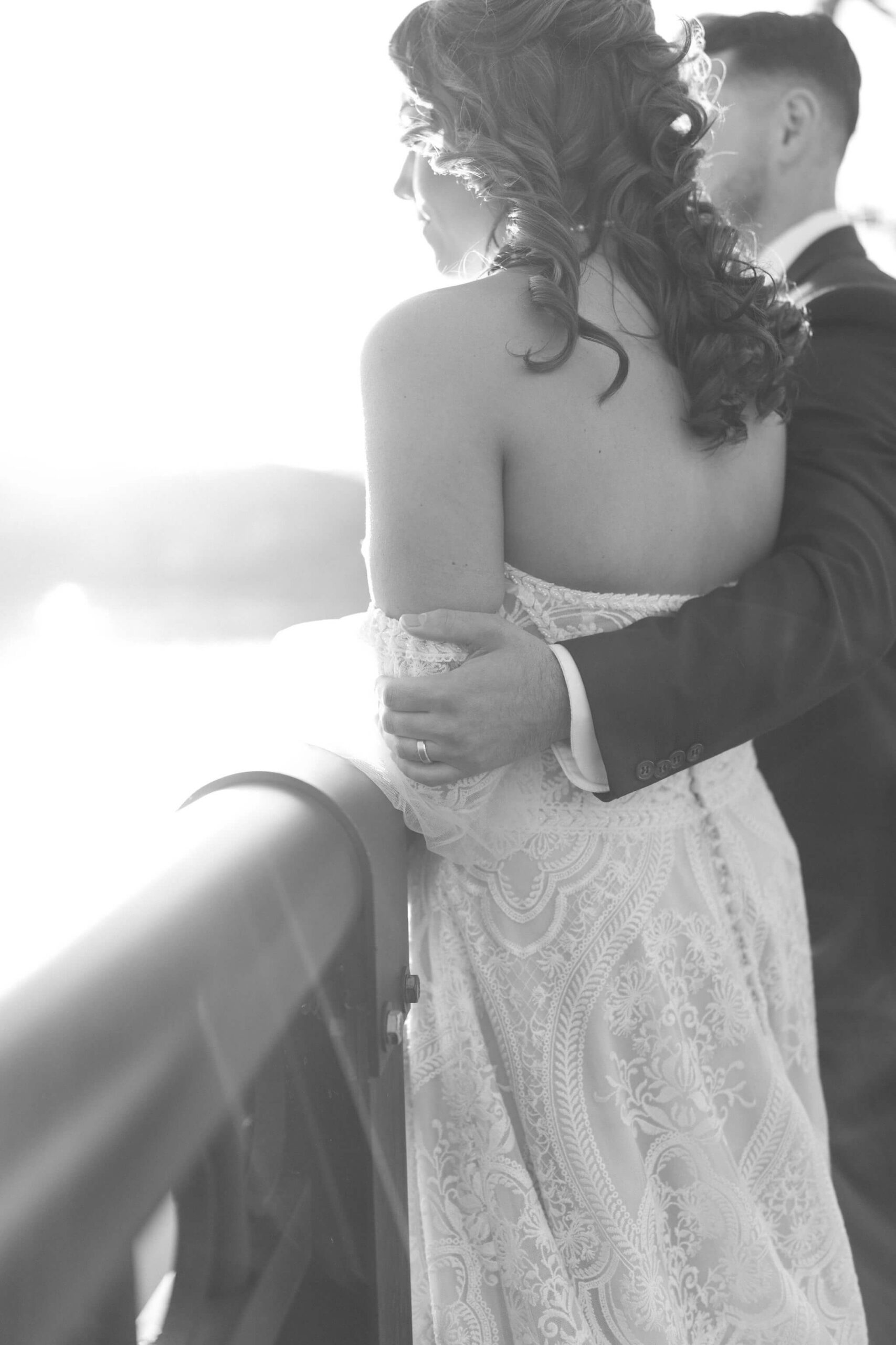 Black and white image of a groom with his arm wrapped around the bride, standing against a railing on a bridge overlooking a river in New Jersey